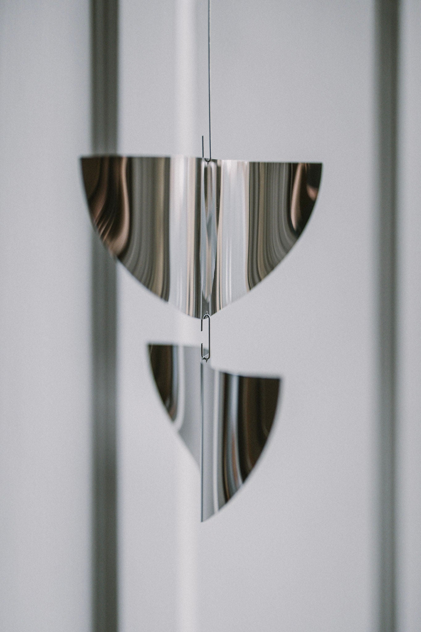 The Sun Catcher by Studio Fie Paarup is a handmade Danish metal mobile with two shiny, semi-circular forms suspended vertically. Its polished steel reflects light beautifully, adding a modern, abstract touch against a blurred gray background.