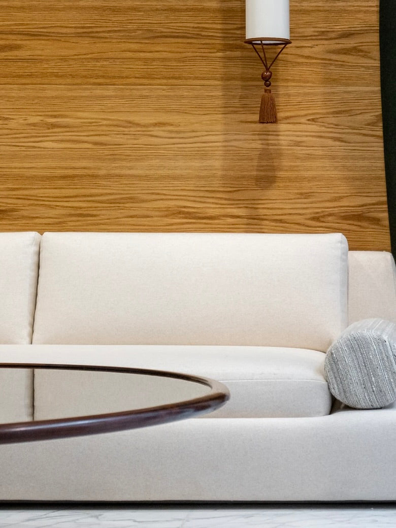 A modern living room showcases the seventies-inspired "Podium" sofa by HAUVETTE & MADANI in washable Dedar cotton velvet set against a wooden panel wall. In the foreground, a round glass coffee table complements the scene, with a cylindrical pillow resting on the sofa. Above, a wall-mounted lamp with a tassel adds to the ambiance.