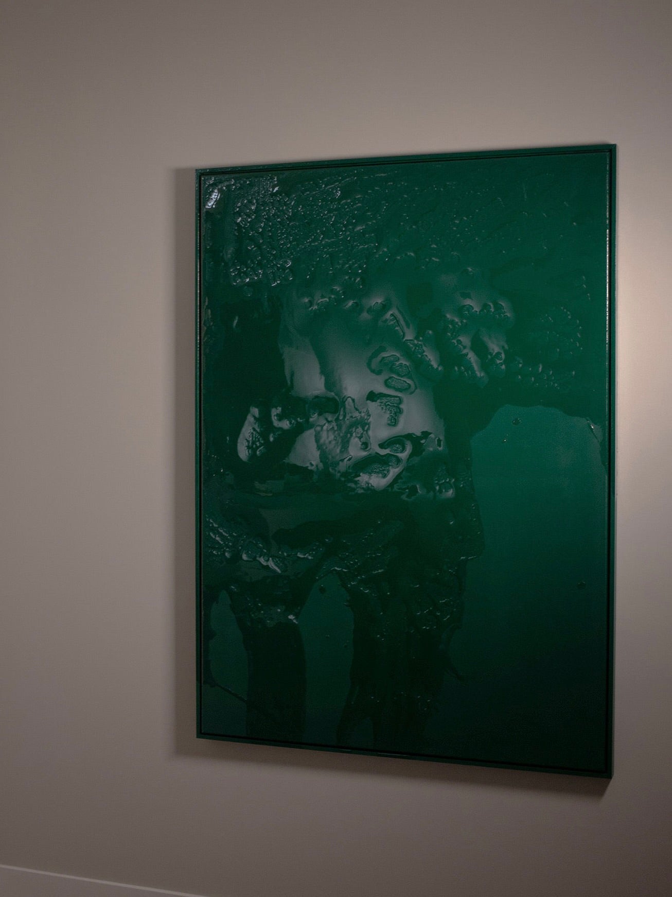 A framed canvas featuring STUDIOPOLS' "Green Flow" painting hangs on a plain beige wall. This dark green abstract artwork showcases textured, fluid-like patterns that evoke a sense of movement across the surface. The deep and intense tones are complemented by shadows that highlight the intricate details of the acrylic paint.