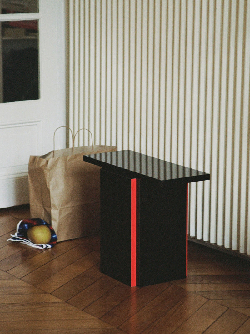 The Mara T Side Table – Red Line by TEGET, with its modern high-gloss black and red accents, stands on a wooden floor near a vertically ridged white wall. Nearby, a brown paper shopping bag and sunglasses showcase stained glass reflections on one lens.