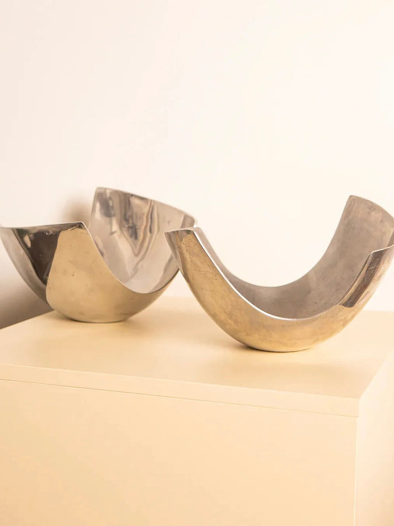 A pair of "U"-shaped fruit baskets from the 80's by Treaptyque, silver-plated with a retro design and shiny, reflective surfaces, rest on a light-colored square pedestal against a plain background. Reminiscent of Italian fruit baskets, one piece is positioned upright while the other is slightly tilted.
