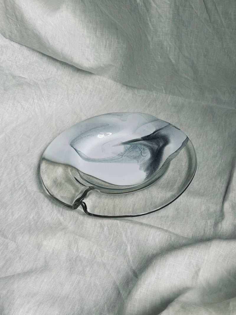 A distinctively-shaped, transparent glass plate with a swirling gray and white pattern from the 1970s Vintage Glass Plate by Hans Jürgen Richartz collection by Flock Studio rests on a soft, light gray fabric background. The texture and design of the plate evoke an artistic and abstract appearance.