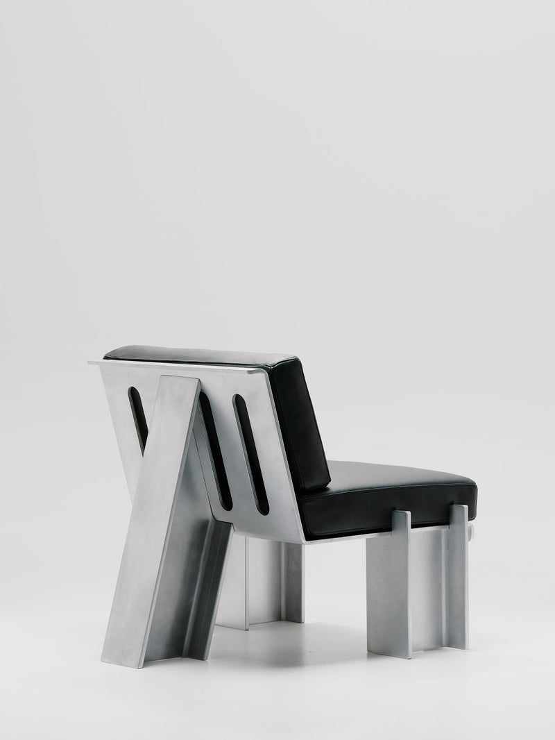 Discover the Beam Lounge Chair by Marquel Williams, featuring a sleek, minimalist design. It includes a waxed aluminum frame and full-grain leather cushions for the seat and backrest. Its angular structure and cutout slots perfectly contrast with a plain, light background.