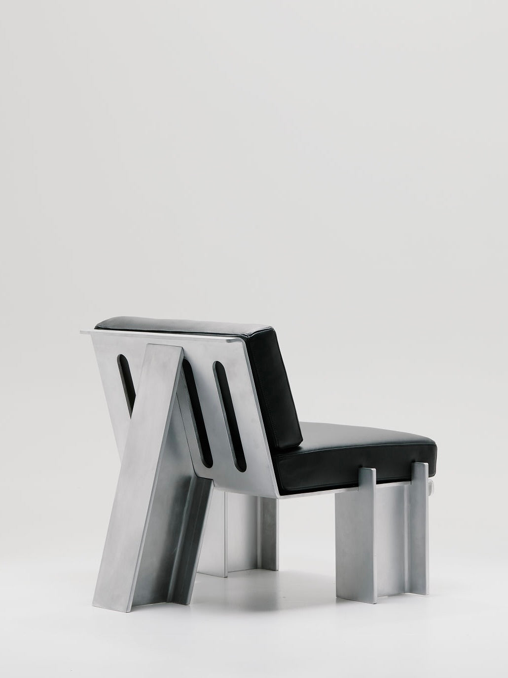 Discover the Beam Lounge Chair by Marquel Williams, featuring a sleek, minimalist design. It includes a waxed aluminum frame and full-grain leather cushions for the seat and backrest. Its angular structure and cutout slots perfectly contrast with a plain, light background.