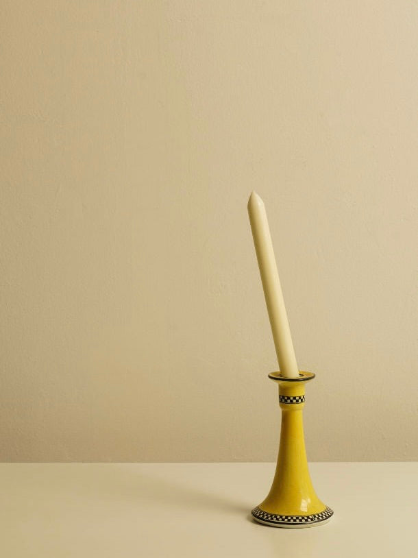 A lit candle stands upright in the Porcelain Candle Holder 1920's by Les Objets Amsterdam, its yellow, intricately designed base placed on a light-colored surface against a beige background. The scene is minimalistic and serene.