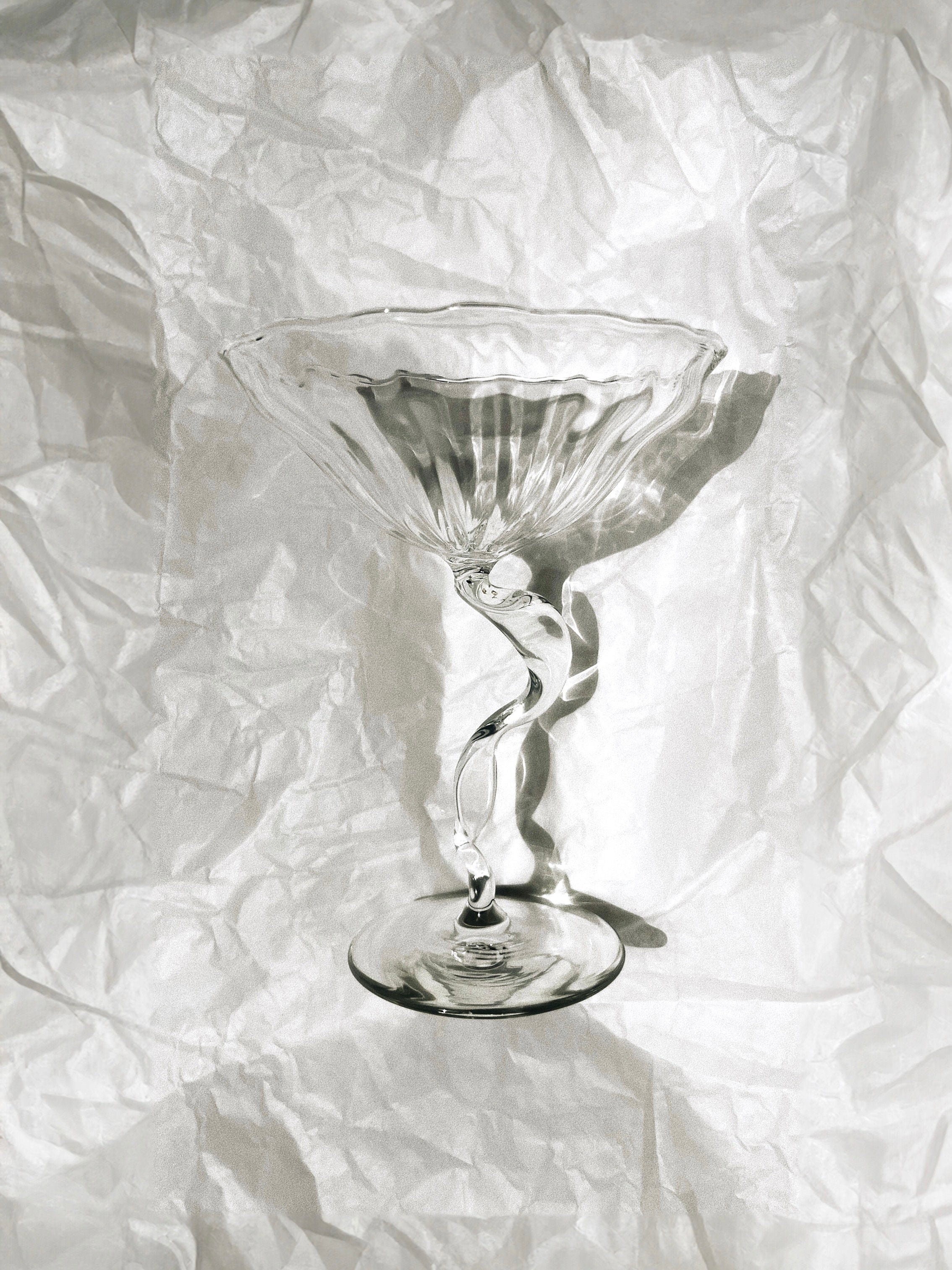 A clear, elegant "Ribbon" Cocktail Coupe with a twisted stem from the Hors-d'Oeuvre collection is placed on a crumpled, textured white surface. Handcrafted in France from borosilicate glass, the light and shadows highlight its exquisite craftsmanship and design.