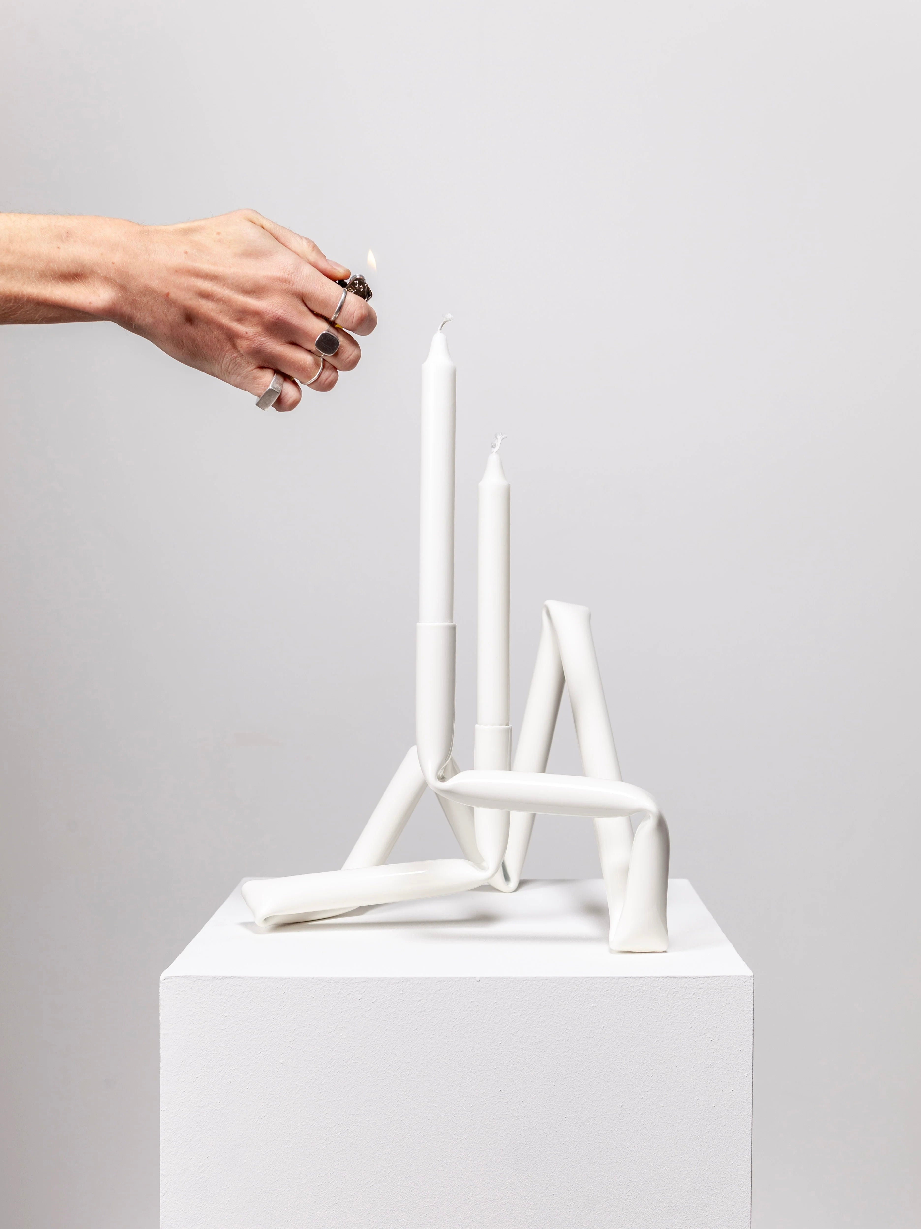 A hand adorned with rings lights a unique Bucatini candle holder from AOT STUDIO's Primi collection. This sculptural white candle holder, featuring three white candles of varying heights, showcases an abstract twisted design made from aluminum tubing and rests on a simple white pedestal against a plain gray background.