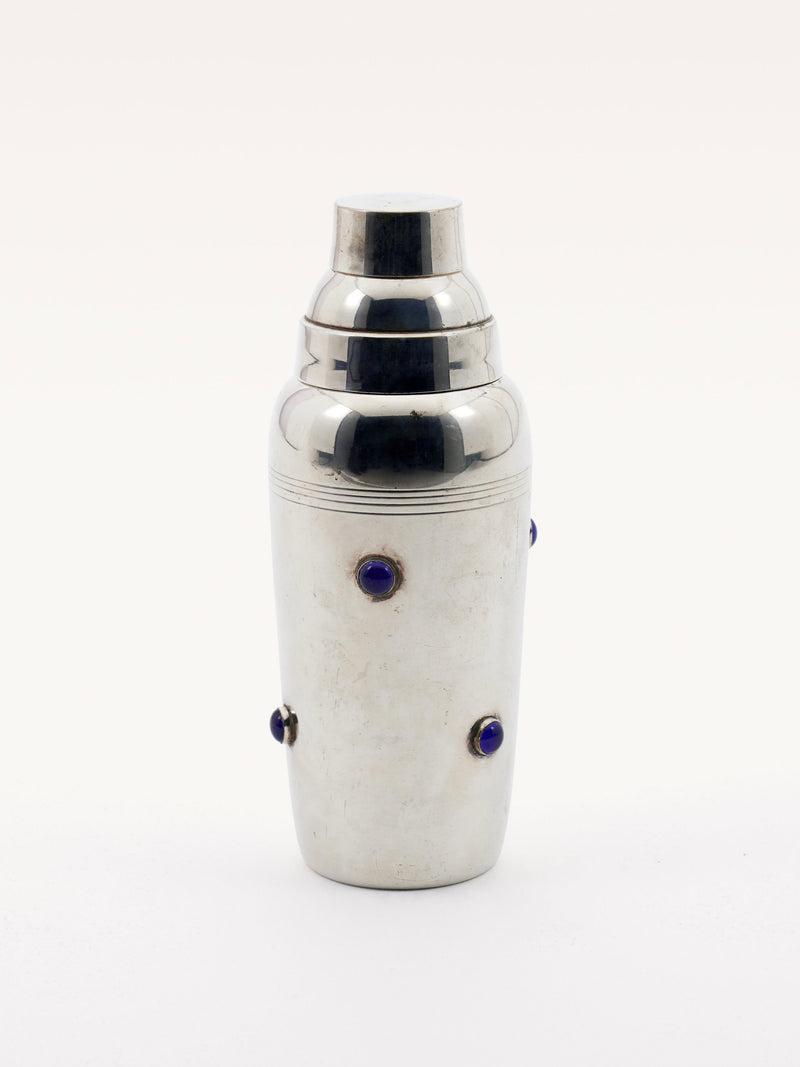 The Shaker with Blue Stones by Galerie MLS features an elegant chrome design, adorned with evenly spaced small blue gemstones. It boasts a smooth reflective finish and a classic, curvaceous shape complete with a detachable lid reminiscent of Cartier style.