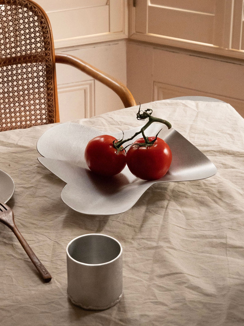 Two red tomatoes sit in the Mid Bowl by Six Dots Design—a modern, curved silver bowl—on a beige tablecloth. A metal cup, a fork, and a woven-back chair are nearby, with sunlight streaming in through North London window shutters.