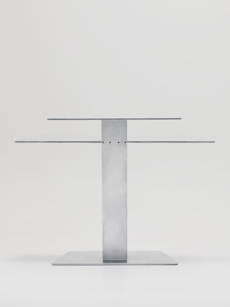 The Beam Desk by Marquel Williams is a sleek, industrial metal table with a minimalist rectangular top and base, connected by a central support. Expertly crafted from laser-cut aluminum, it embodies architectural design principles in a plain white backdrop.