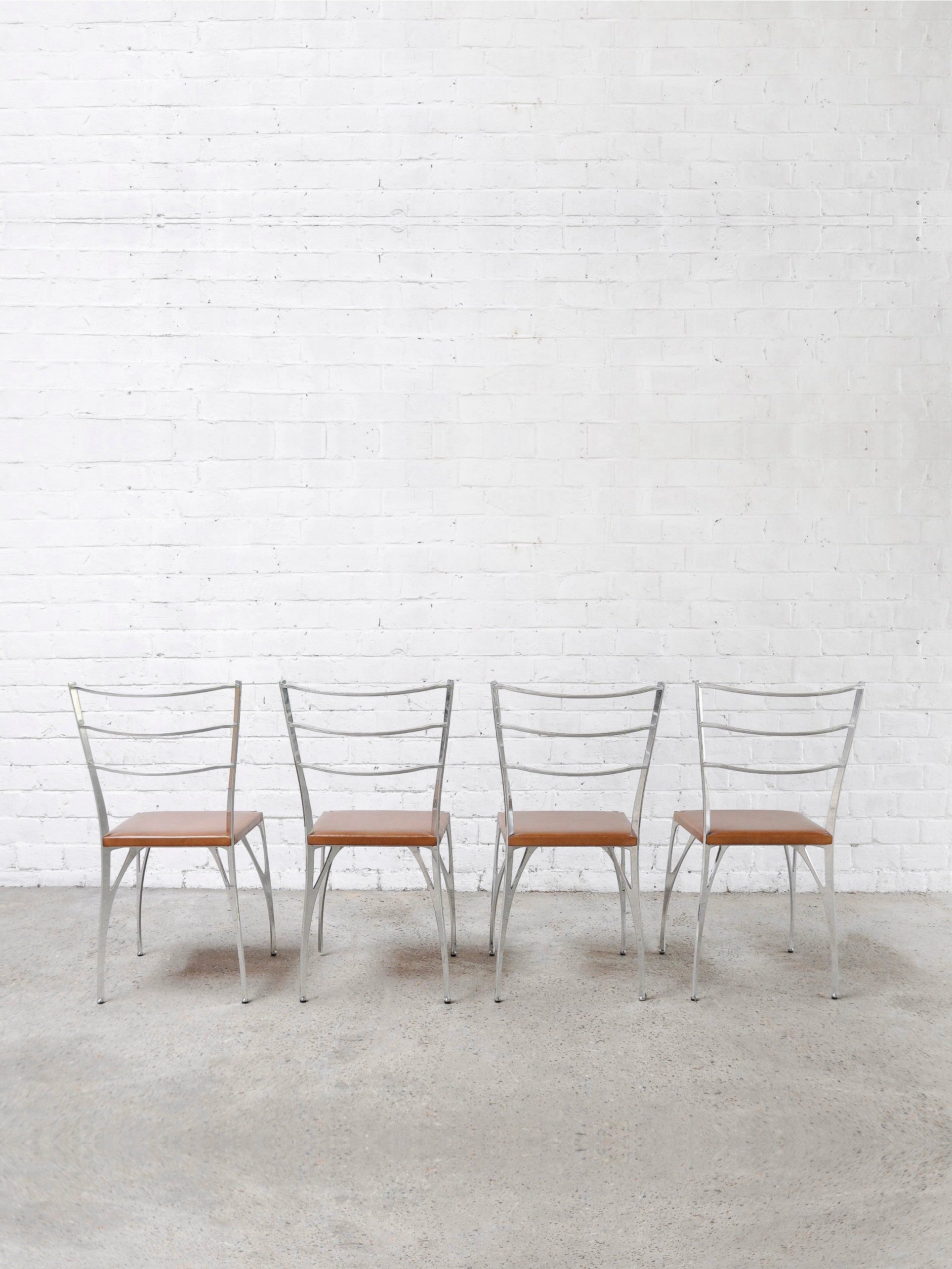 A set of four 'Organic' dining chairs by Erwan Boulloud, featuring sculptural aluminum frames and wooden seats, are lined up against a white brick wall. The light-colored, textured concrete floor enhances their artful presence. Brand: Introverso.