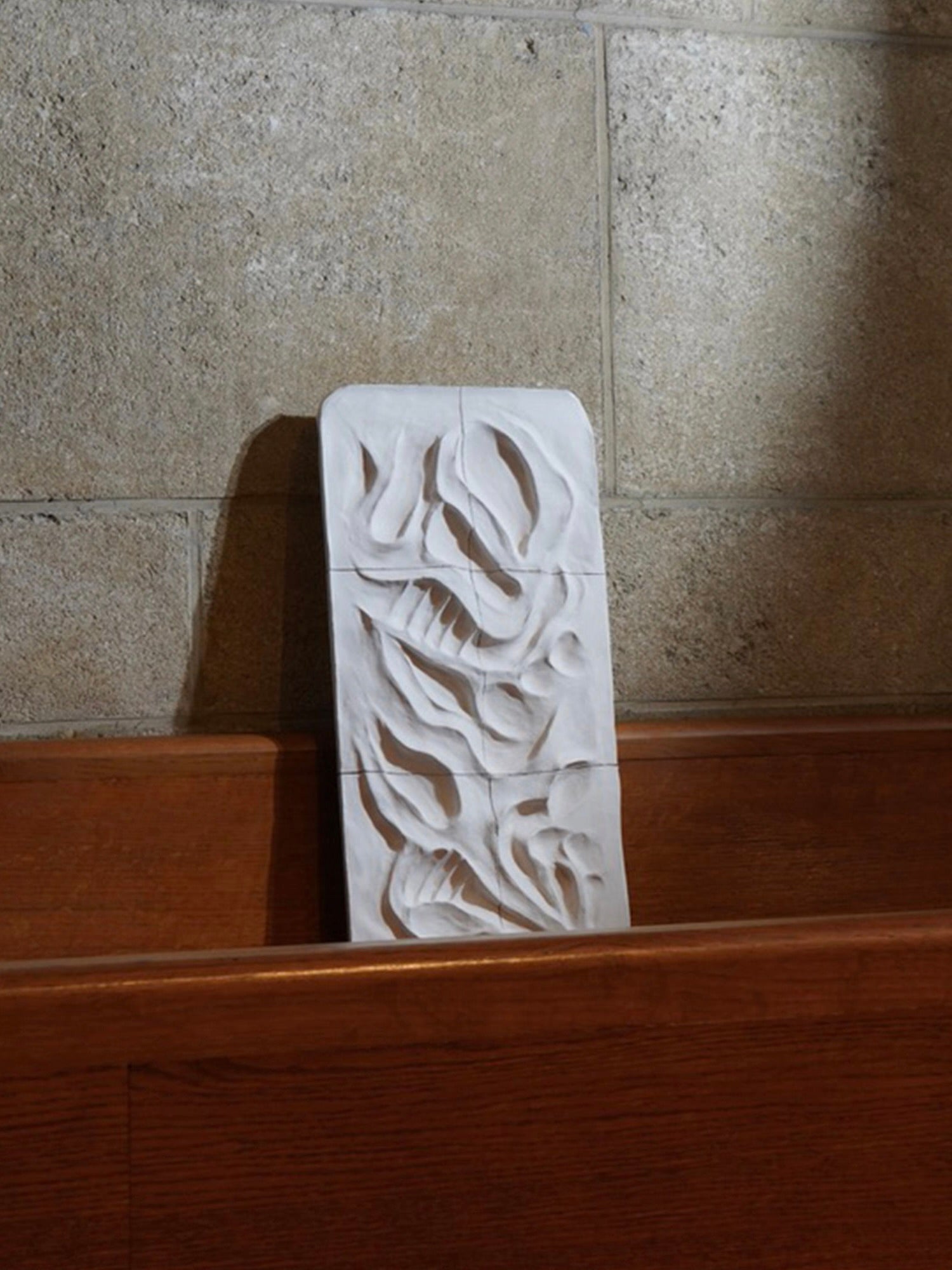 The Death Valley - Wall Sculpture by Franck Scala, a white ceramic bas-relief with abstract floral designs, leans against a textured stone wall atop wooden pews, evoking the serenity of a chapel.