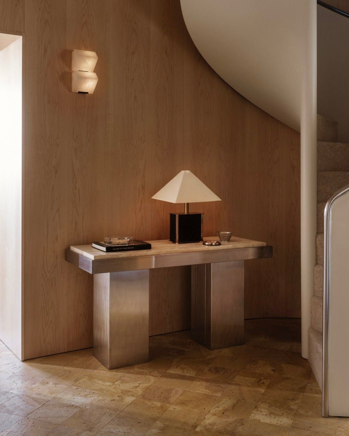 A modern interior featuring a wooden wall and a spiraling staircase showcases the Console Table 01 by Studio Vraco, crafted in brushed stainless steel, supporting a square-based lamp. The tiled floor leads to a glimpse of the dining area adorned with wall art and a patterned rug through the doorway.