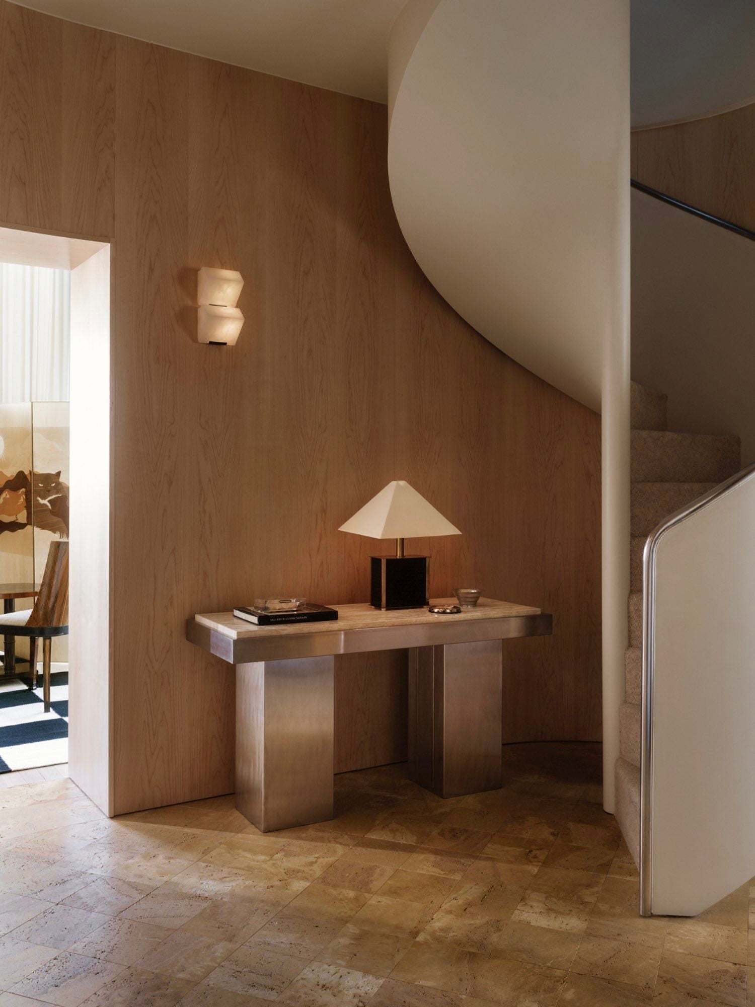 A modern interior featuring a wooden wall and a spiraling staircase showcases the Console Table 01 by Studio Vraco, crafted in brushed stainless steel, supporting a square-based lamp. The tiled floor leads to a glimpse of the dining area adorned with wall art and a patterned rug through the doorway.