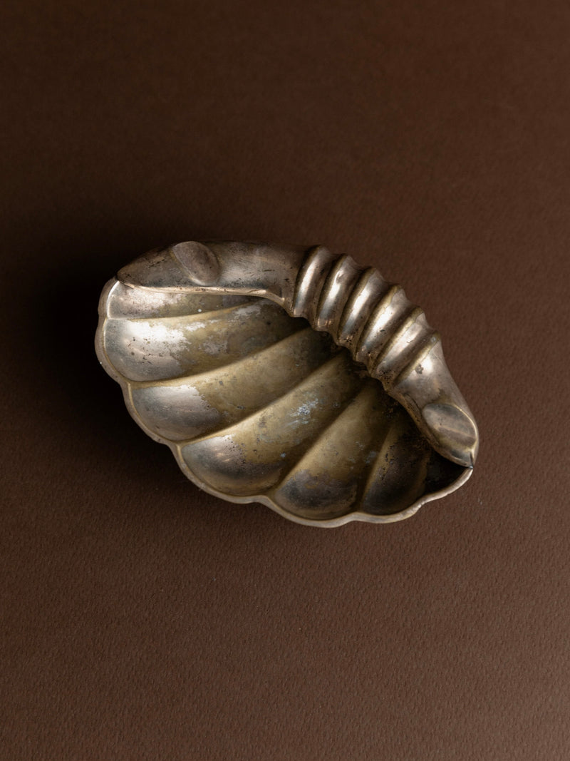 An Ashtray in Shell Shape from Bottega Jacobs, dating back to the 1960s in Italy, graces a brown surface. It features a ribbed, curved handle and boasts a tarnished patina that accentuates its intricate details and texture.