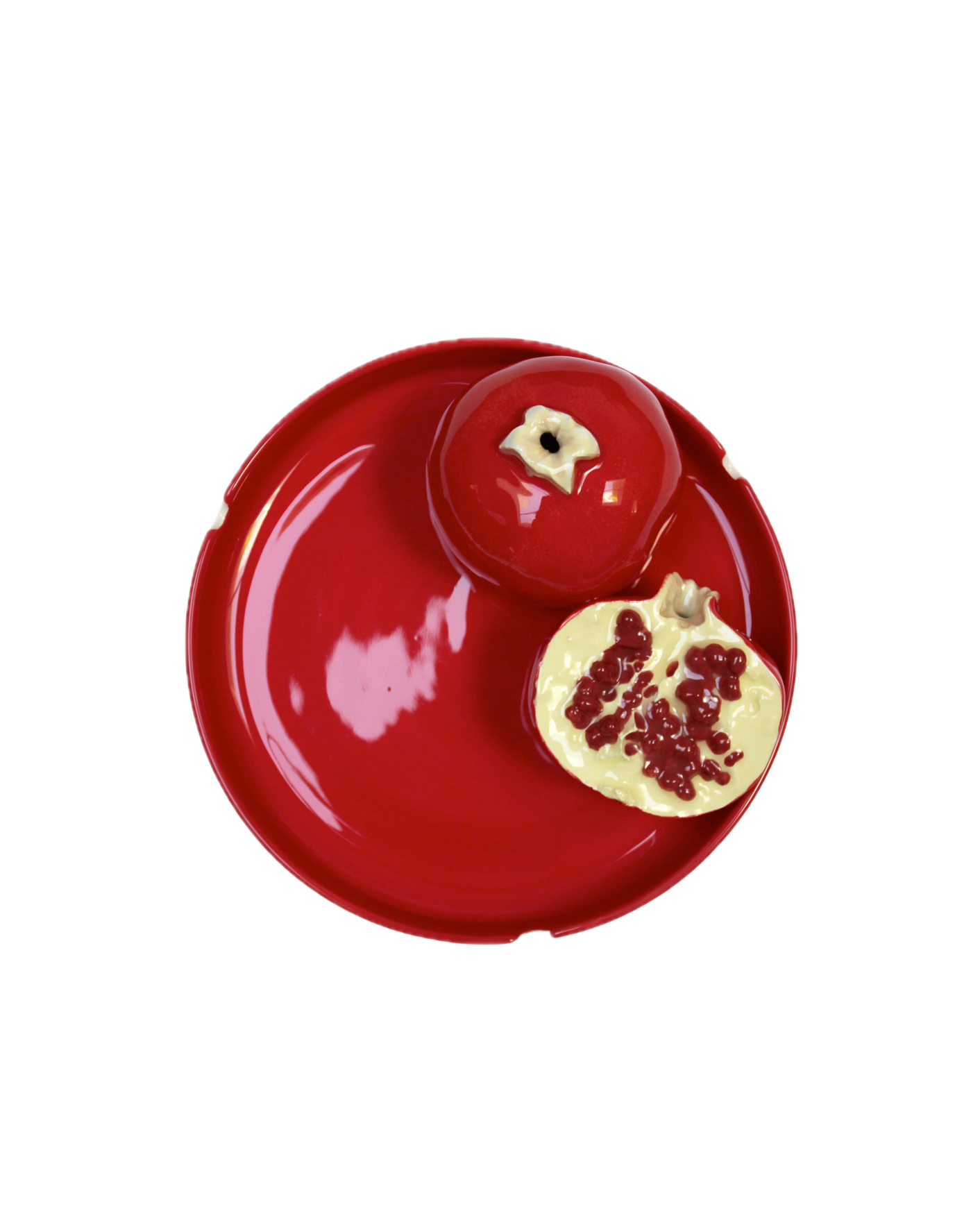 A vibrant red plate from Villa Arev holds a ceramic Grenades Ashtray. One ashtray is whole, while the other is sliced open, revealing detailed seeds inside. The glossy red of the painted earthenware creates a striking, cohesive look infused with Mediterranean charm.