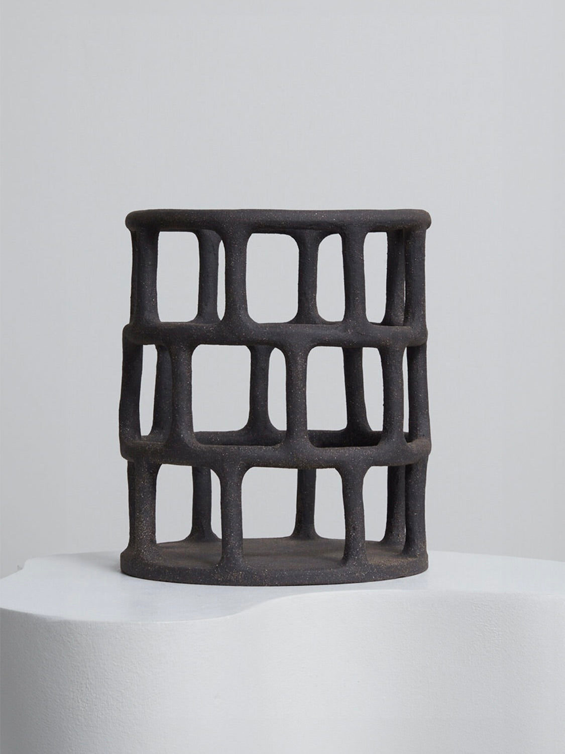 The Handcrafted Black Basket by Solenne Belloir, featuring a dark cylindrical open-grid design with a rough texture, rests on a smooth white surface against a light gray background.