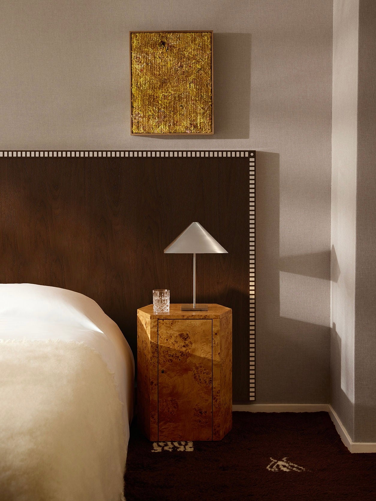 A cozy bedroom features a bed with white bedding, a dark wooden headboard, and a meticulously crafted nightstand from Lemon, called the Pair of Park Night Stands in Poplar Burl. The nightstand hosts a modern lamp with a conical shade and a glass of water. Above the headboard is textured, gold-toned artwork that complements the room's warm lighting.