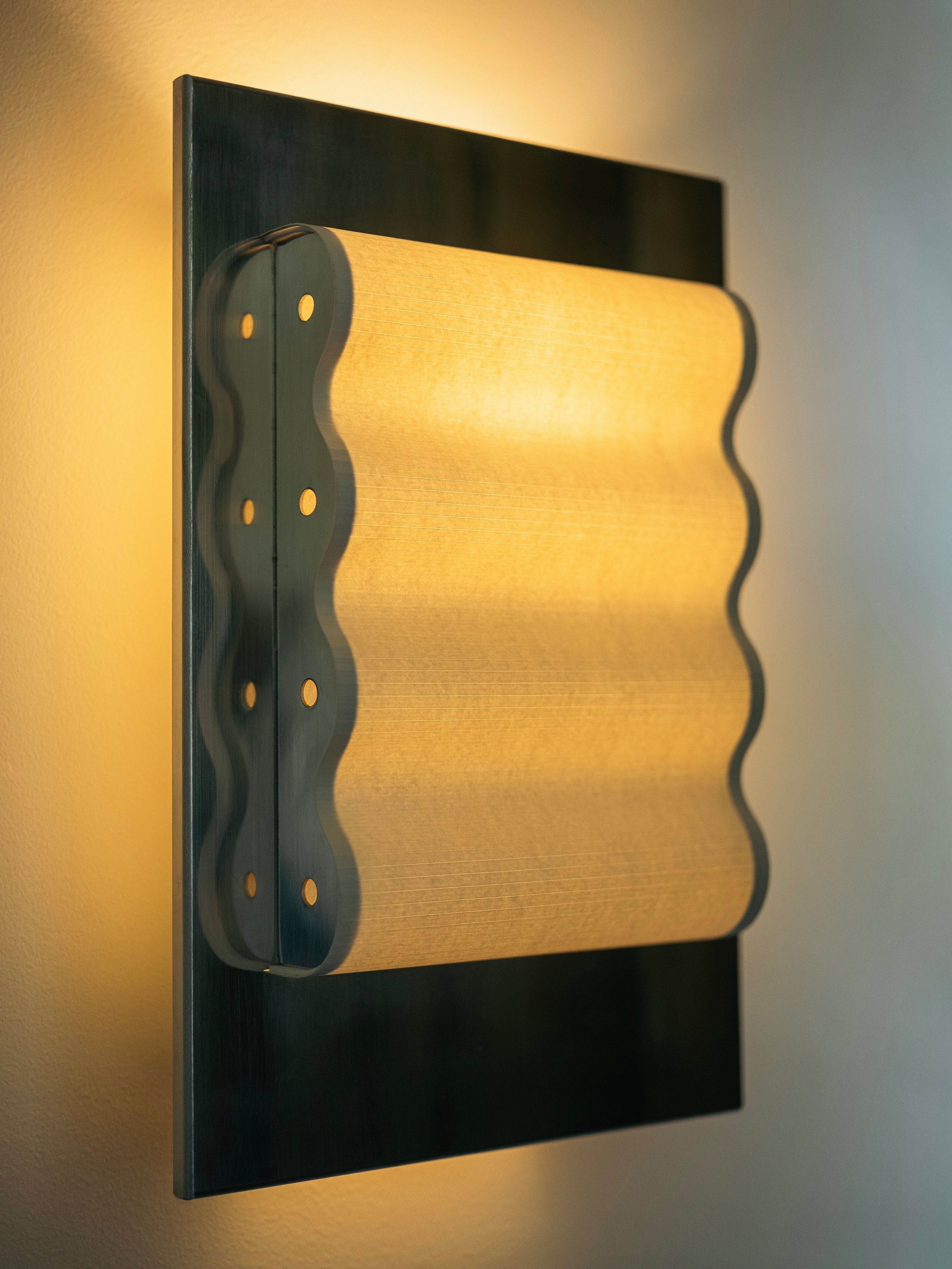 The Wall Light Frame Miroir S by Violaine d'Harcourt emits a warm, ambient glow against a light-colored wall. The lamp features a rectangular metal plate with a wavy, perforated sheet of frosted glass or plastic that diffuses the light. Its design combines sleek metal and soft curves for a minimalist aesthetic that echoes modernist architecture.