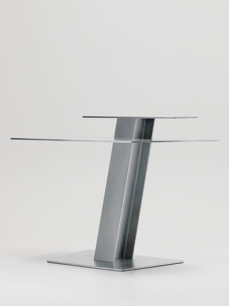 The Beam Desk by Marquel Williams is a minimalist steel table base with a geometric design and handcrafted touch. It has two horizontal steel plates, a central slanted beam, and a rectangular base, shown against a plain light gray background.