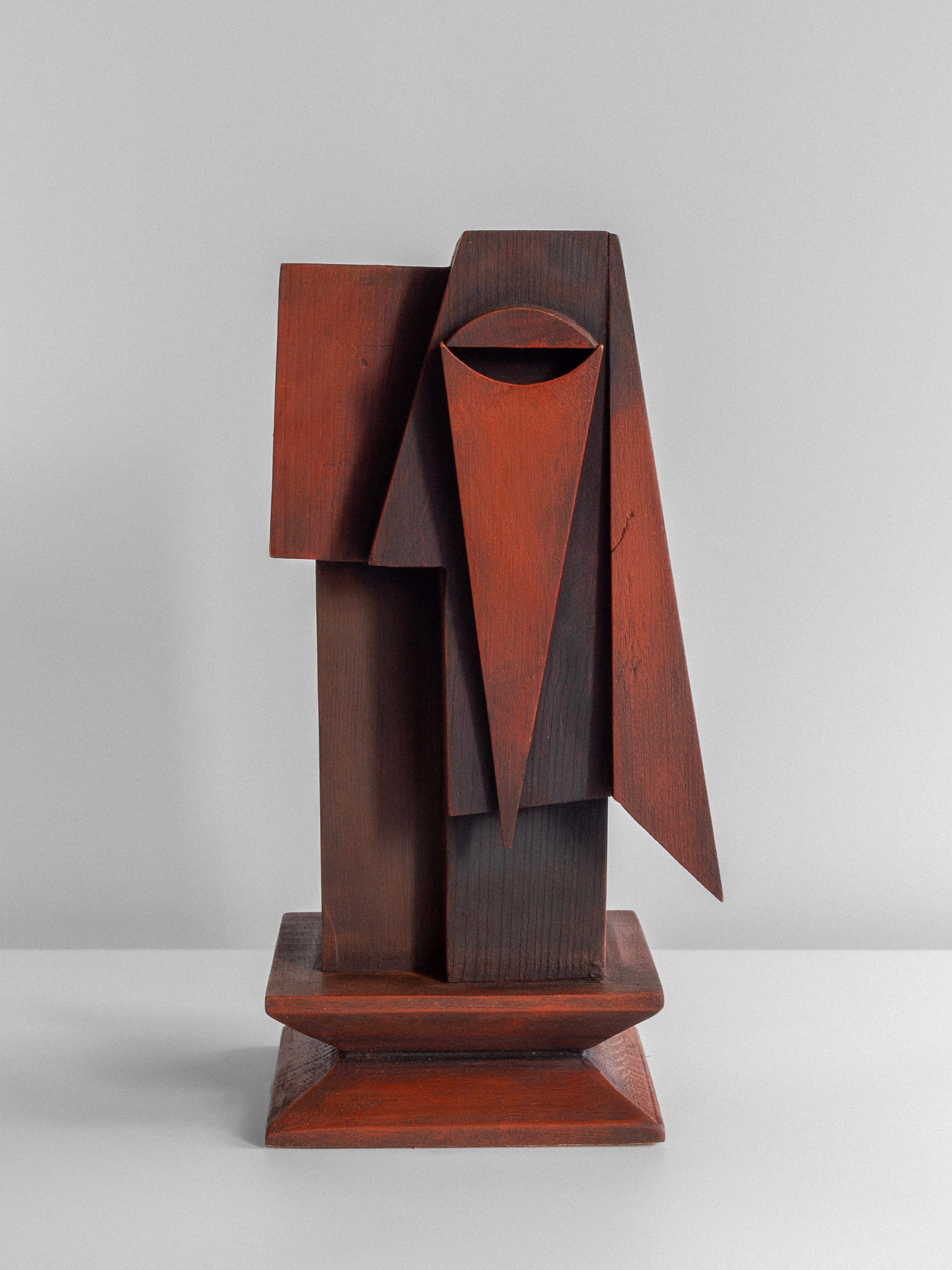 The Vintage Cubist Sculpture by Atelier Boulogne from Spigel is a figural wood piece inspired by cubism, featuring geometric shapes like triangles and rectangles. The abstract design forms a stylized face with a long, triangular nose and a single half-oval eye. This reddish-brown sculpture is beautifully displayed on a flat white surface.