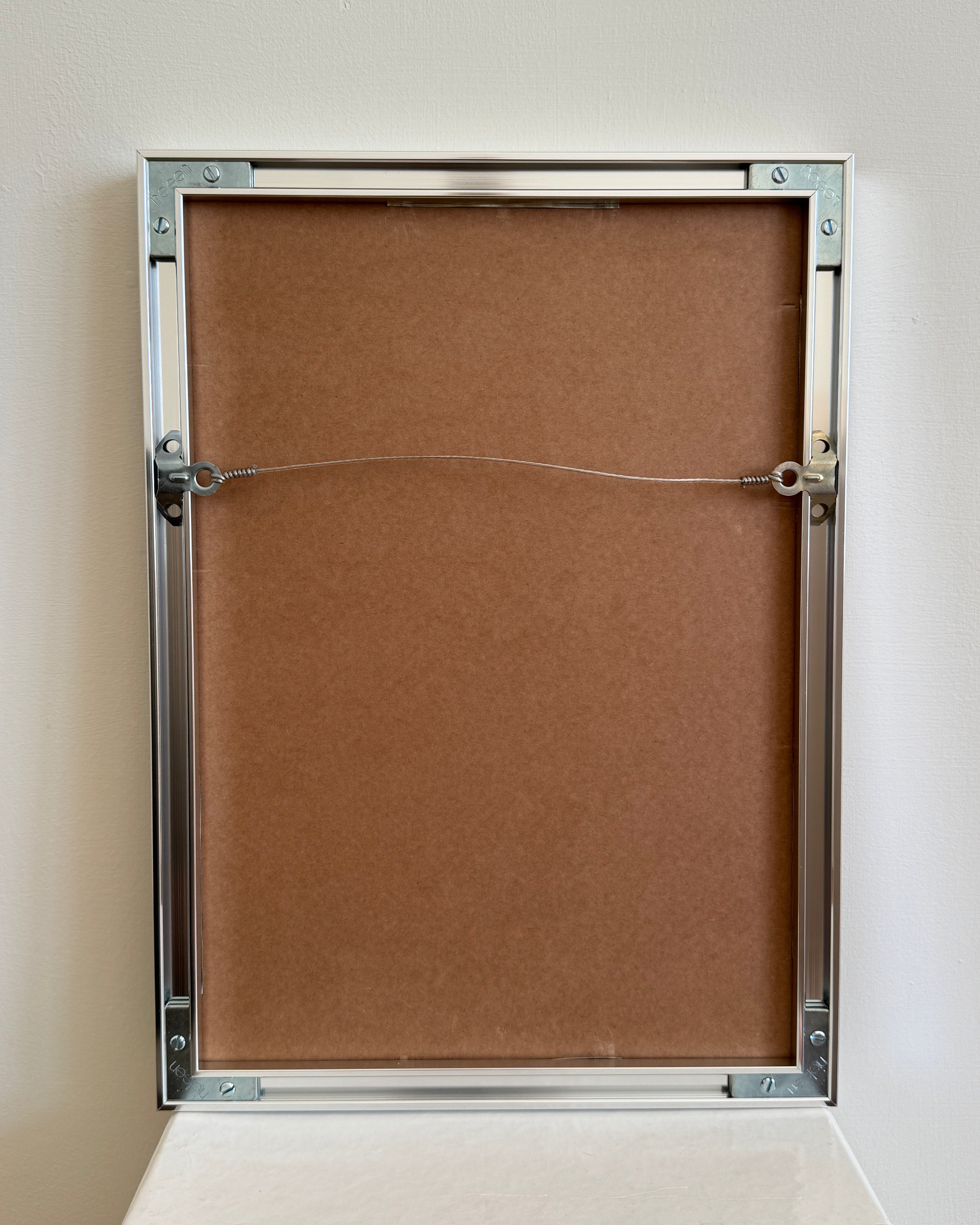 A brown-backed picture frame with a silver metallic border is shown from the rear. The frame holds Villa Necchi Campiglio, Milan - Print Photography by Grace Rose Watts, has a hanging wire on side mounts, and rests against a plain white wall.
