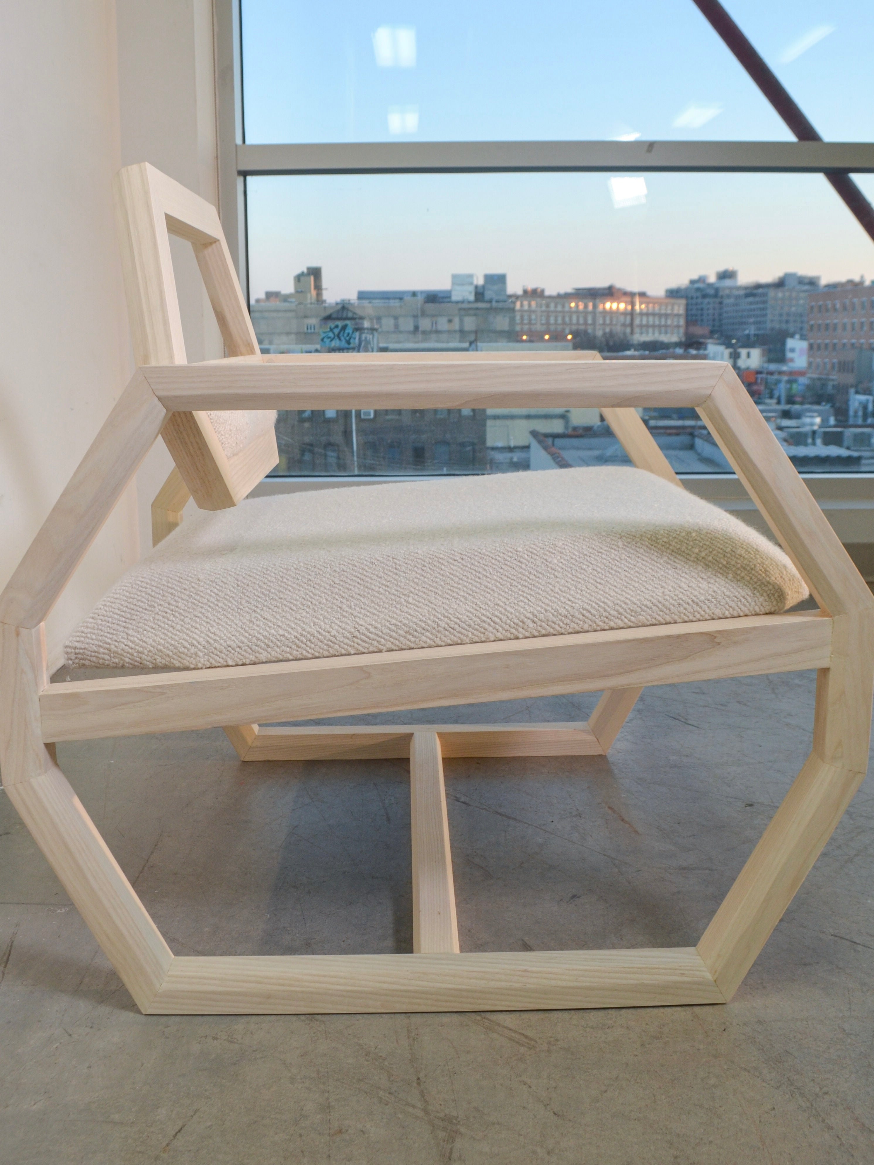 A modern Marcel Accent Chair by Nathan Chintala Design LLC, featuring a geometric hexagonal frame and a cream-colored cushioned seat, is positioned in front of a large window. Beyond the window lies an urban cityscape, complete with buildings standing tall under a dusky sky.