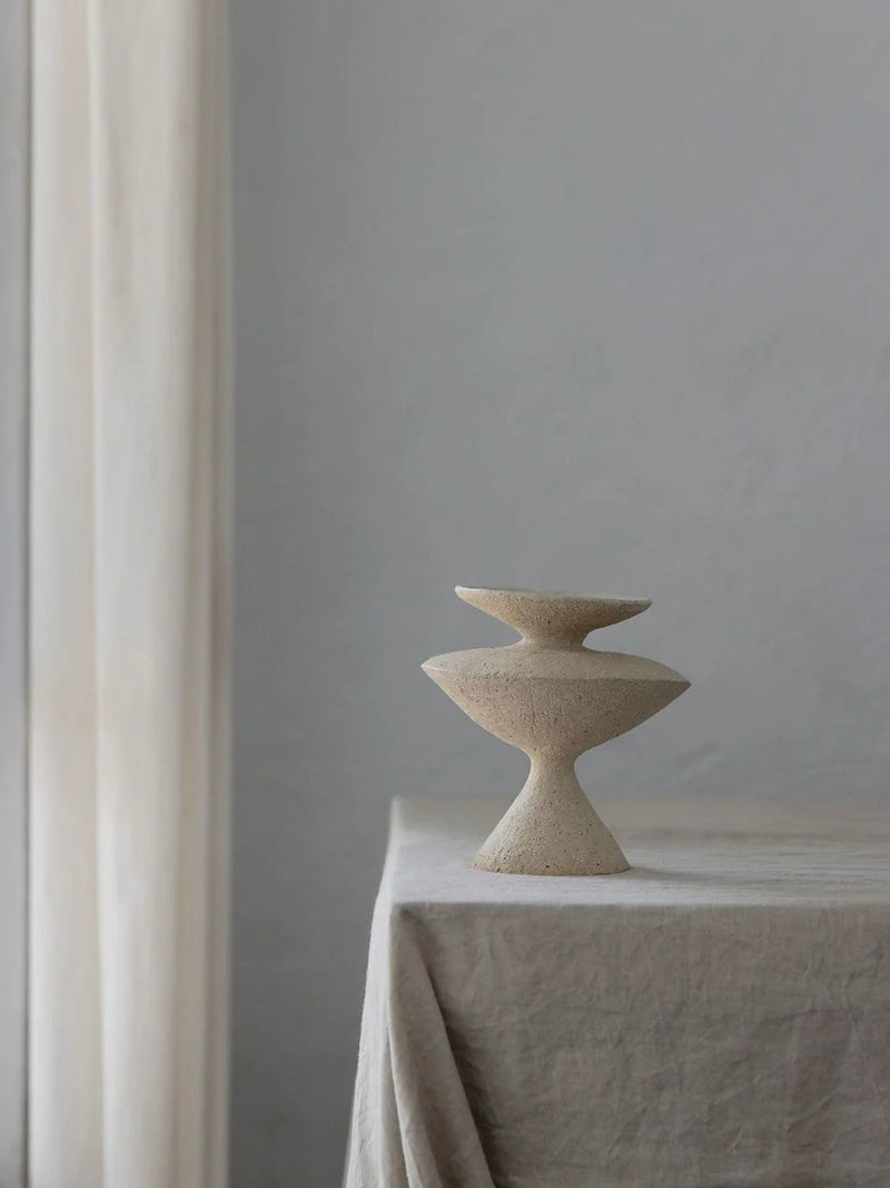 The Curve Object by Emma Lindegaard Studio, a minimalist ceramic sculpture crafted with the coiling method and featuring geometric shapes, sits on a linen-covered table. Soft natural light filters through sheer curtains, casting gentle shadows and highlighting its textural beauty.