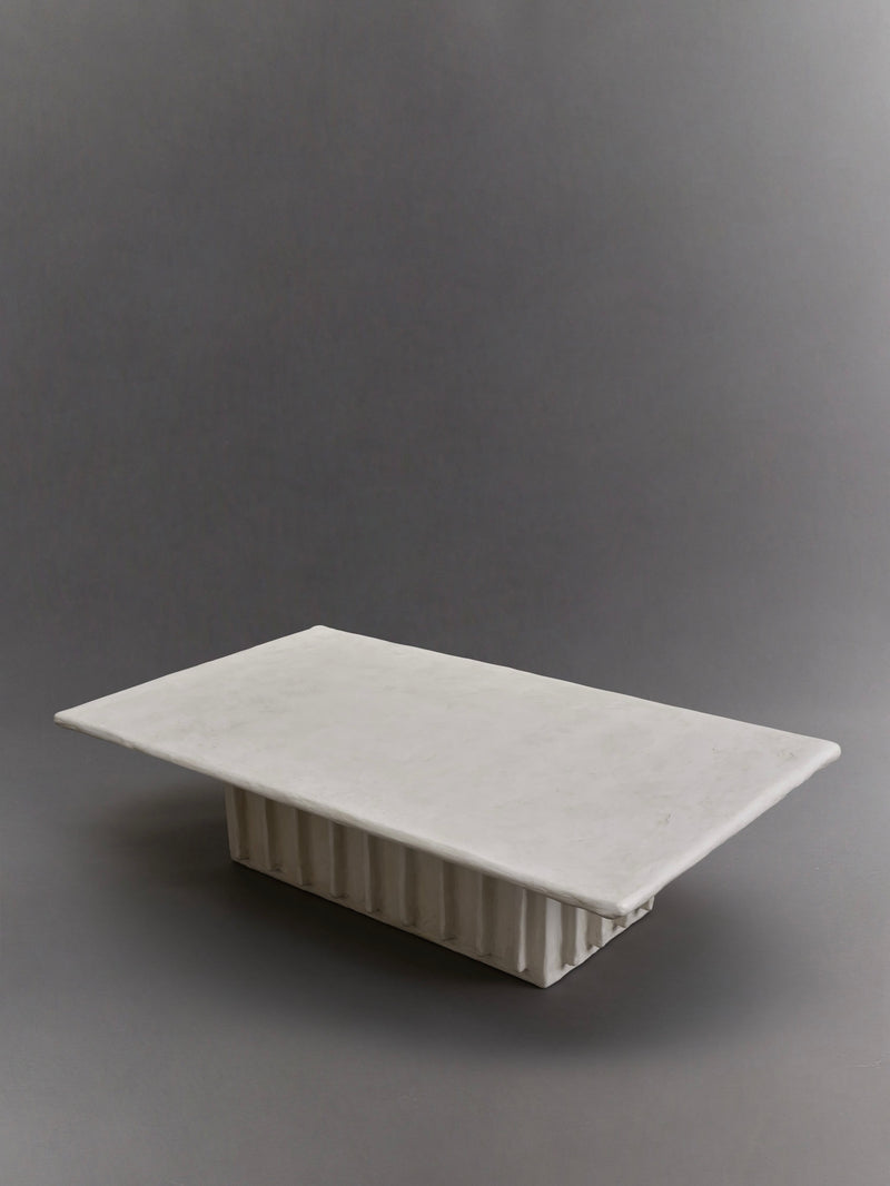 The Columnar Coffee Table by Ombia Studio is a minimalist, rectangular marble piece featuring a smooth top and ribbed base against a plain gray background. This unique, made-to-order item can be customized to fit your space perfectly.