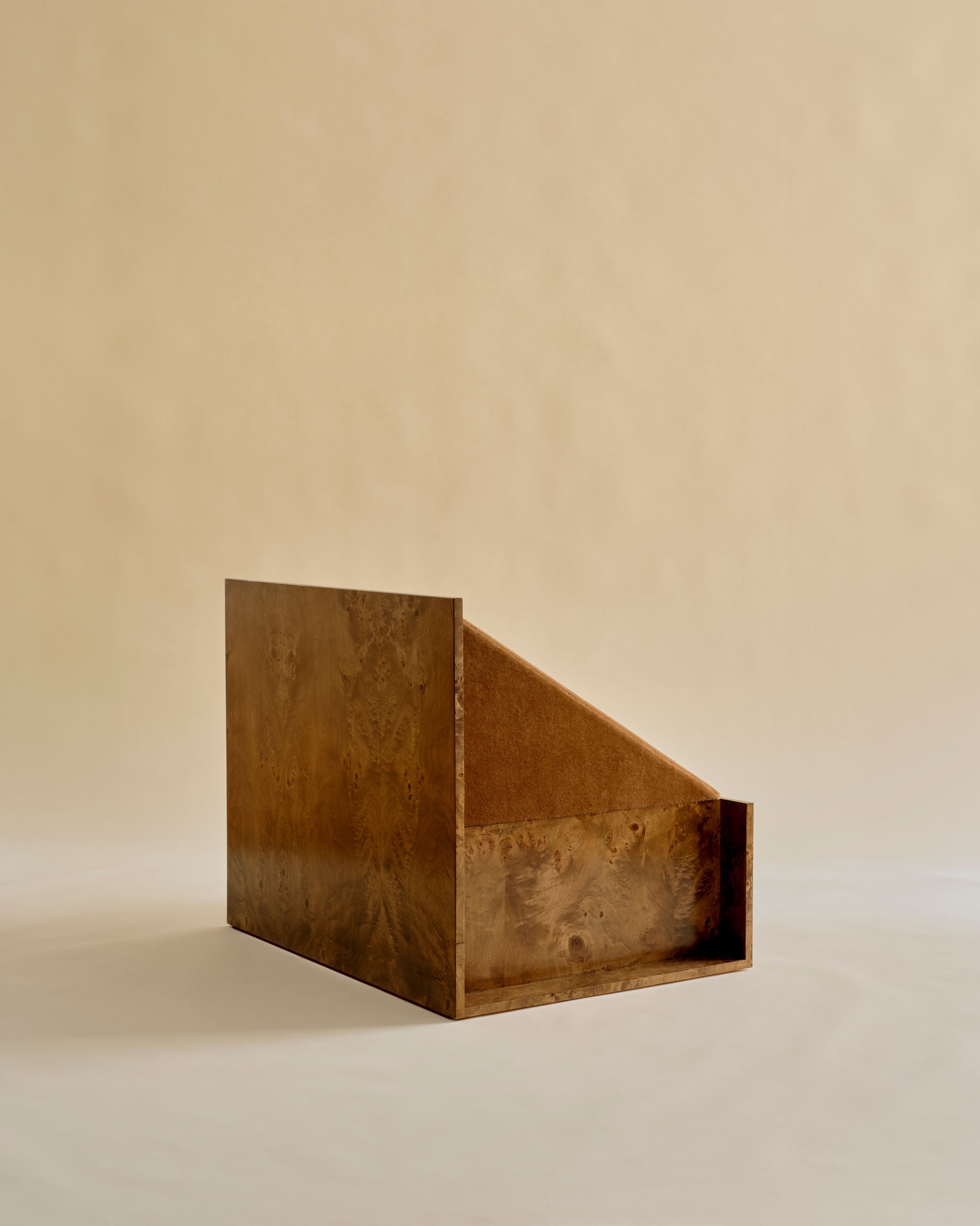 A wooden object with a rectangular base and slanted side, reflecting the minimalist elegance of Known Work's 'Perceptions' Armchair, stands against a plain beige background resembling a refined magazine or book holder.