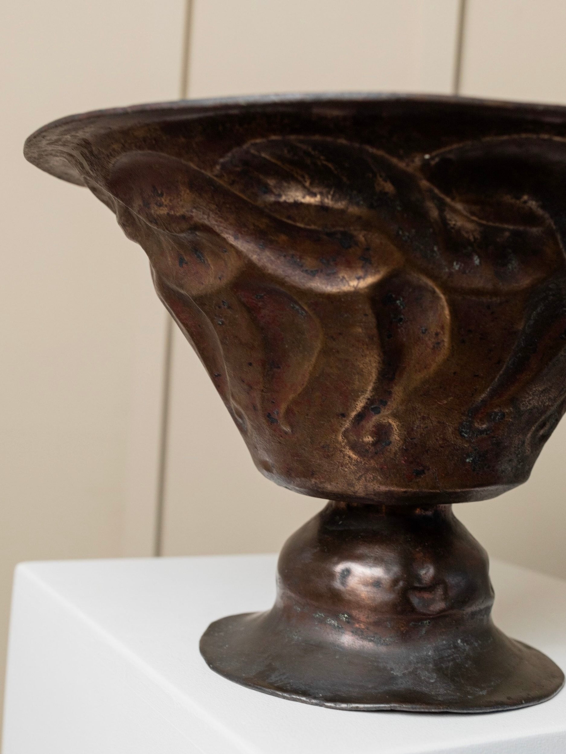 A close-up of the Antique Fruit Bowl - Hand-Carved Copper by Bottega Jacobs, featuring an ornate, bronze-like textured wavy design with a subtle antique patina. The fruit bowl is displayed on a white pedestal with a soft-focus beige background.