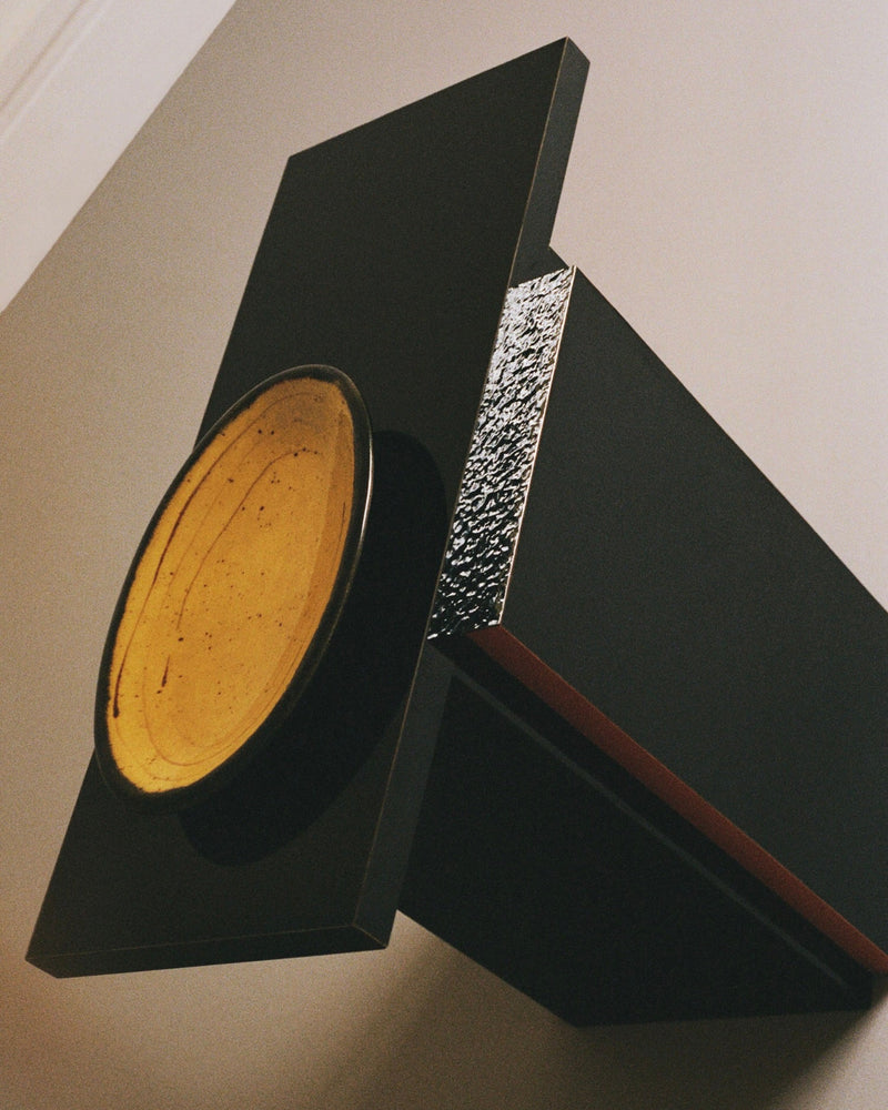 The Mara T Side Table – Red Line by TEGET features a high-gloss black geometric design with metallic textures. A round yellow disk contrasts against the dark structure, resembling sophisticated laminated furniture, and is mounted on a wall for a striking modern art piece effect.