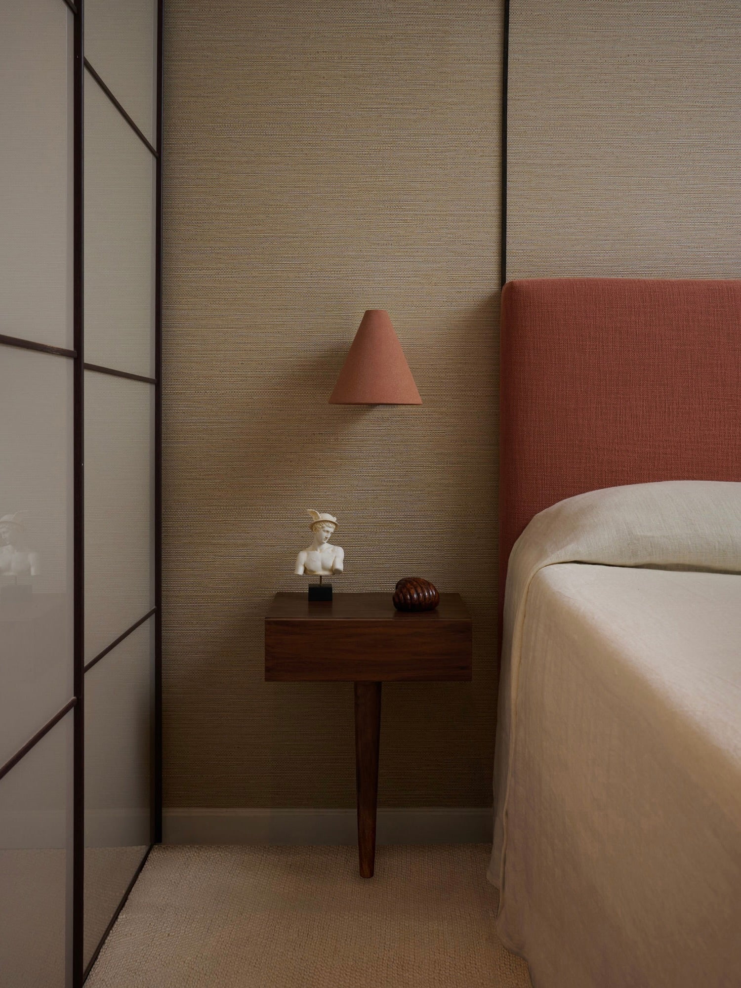 A minimalist bedroom showcases a pink upholstered headboard against a brown accent wall. A wooden bedside table holds a small stoneware sculpture for decoration. Above, the 'Esoteric' Almond Brown Wall Light by Mariza Galani casts a warm glow, while neutral-toned bedding enhances the serene ambiance.