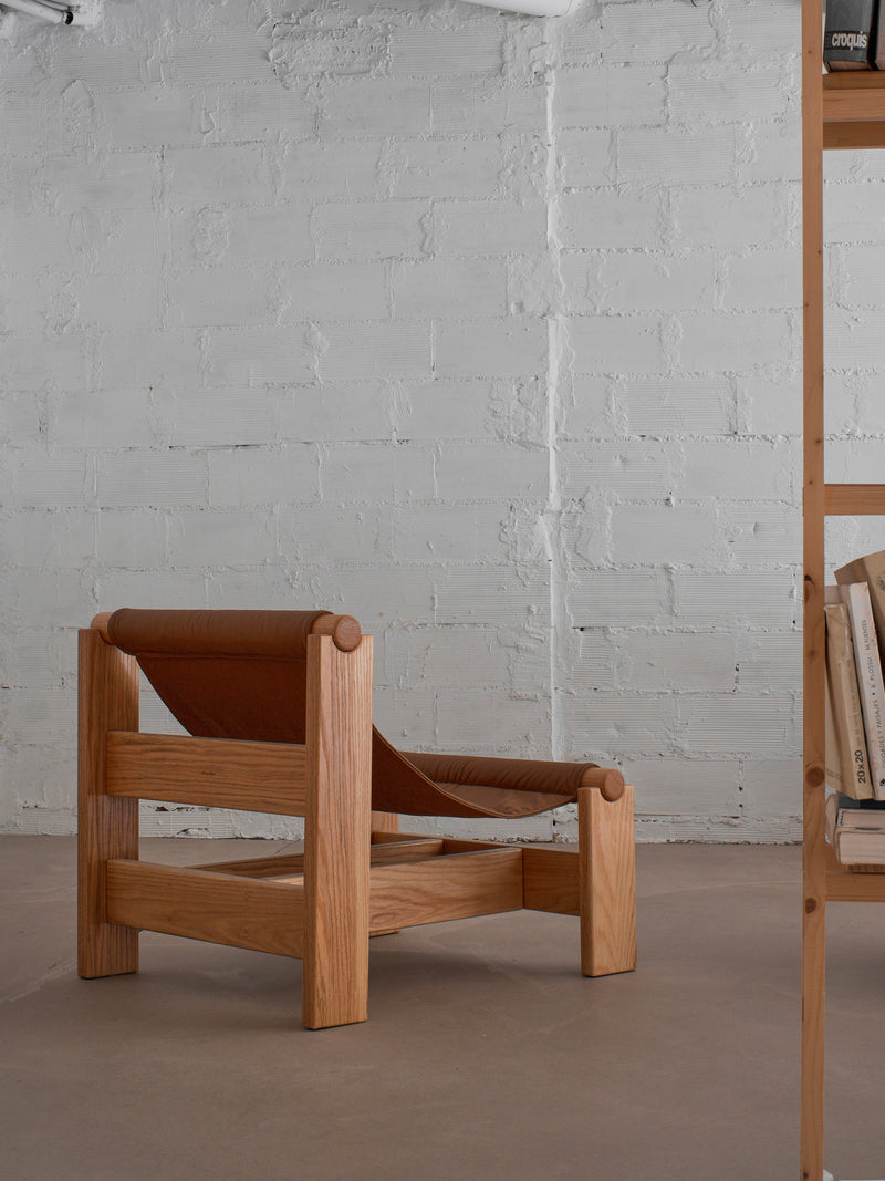 The malva "Lounge Up" Lounge Chair in brown leather stands in a room with light gray brick walls and a beige floor. A wooden bookshelf with books appears partially on the right.