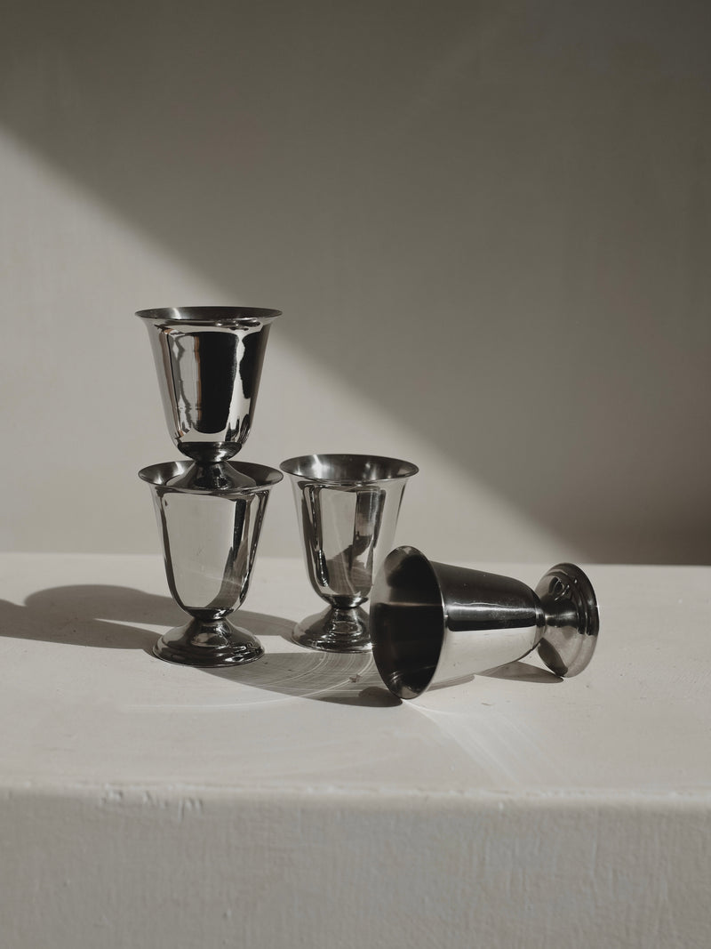 A Set of 4 Coupes by Guy Degrenne from septembre studios is displayed on a white surface: three are upright and stacked, the fourth lies on its side. Soft lighting emphasizes their classic 1970s stainless steel design.