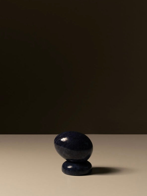 The Marble Egg Sculpture by Les Objets Amsterdam, crafted from dark, smooth lapis marble, sits atop a small round base against a minimalist, dark brown background. Soft lighting casts a gentle shadow on the flat surface below the stone, imbuing this elegant decoration piece with subtle grace and symbolic significance.