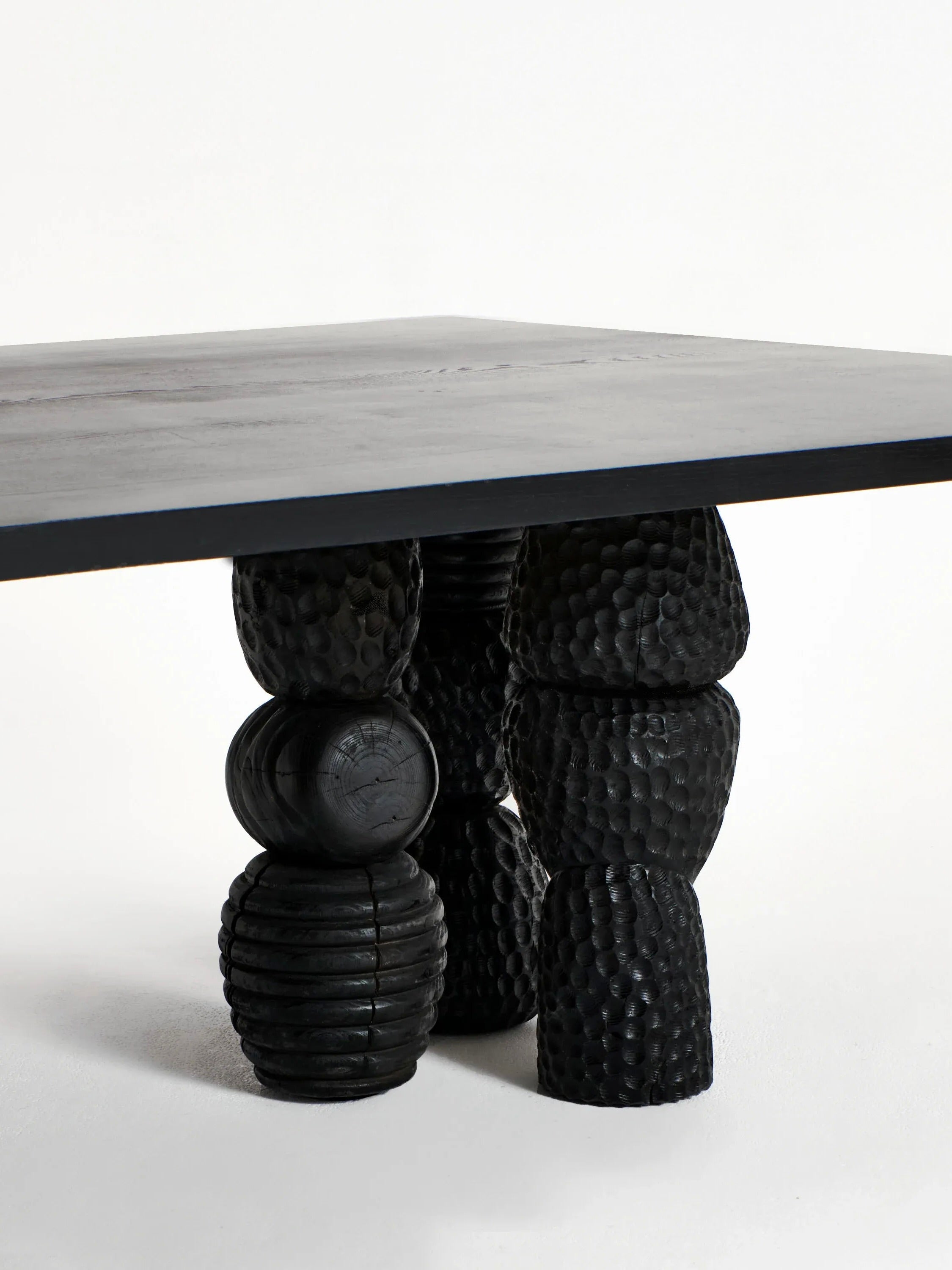 A detailed view of the Menina Dining Table from Project 213A showcases a sleek black surface paired with intricately textured, sculptural legs crafted from chestnut wood. These legs feature stacked, rounded forms with a hammered-like finish, highlighting the handcrafted nature of this contemporary and artistic design.