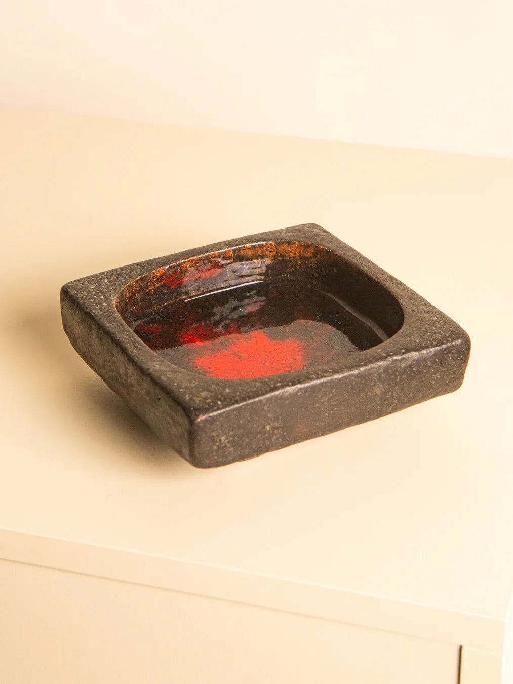 Set against a light-colored backdrop, the 60's Square Ceramic Tray by Jan Van Erp from Treaptyque displays its square black ceramic form with a striking red glazed inner surface. This decorative piece exudes a rustic, handmade charm, seamlessly blending functionality and elegance into one unique work of art.