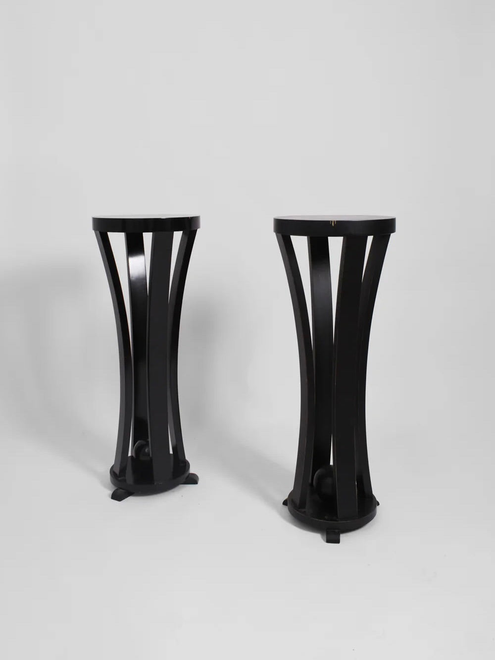 A pair of black wooden pedestals from the 1960s, offered by Barracuda Interiors, features a modern cylindrical design with curved legs and is set against a plain white background. Their sleek, symmetrical style is reminiscent of 1960s France as they stand side by side.