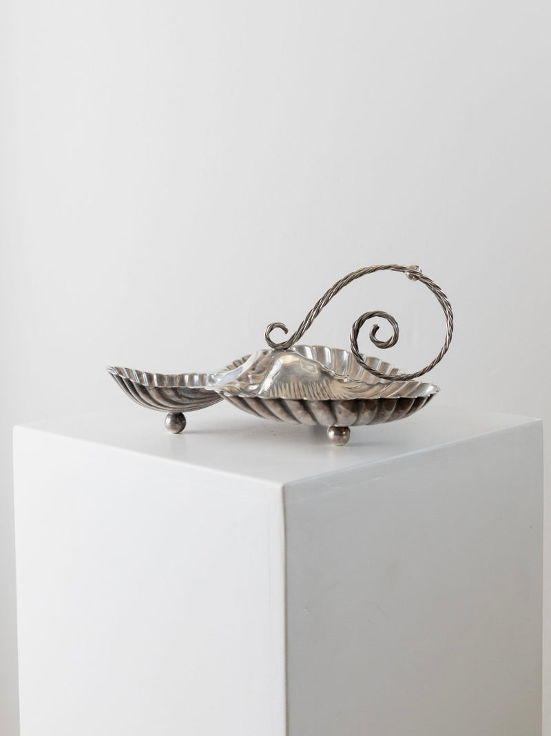 The Silverplated Centrepiece Shell Shape by Bottega Jacobs rests on a white pedestal. This vintage centerpiece showcases a leaf-like design with an ornate swirling handle, featuring a ridged texture and standing on four small spherical feet, set against a plain, light-colored wall.