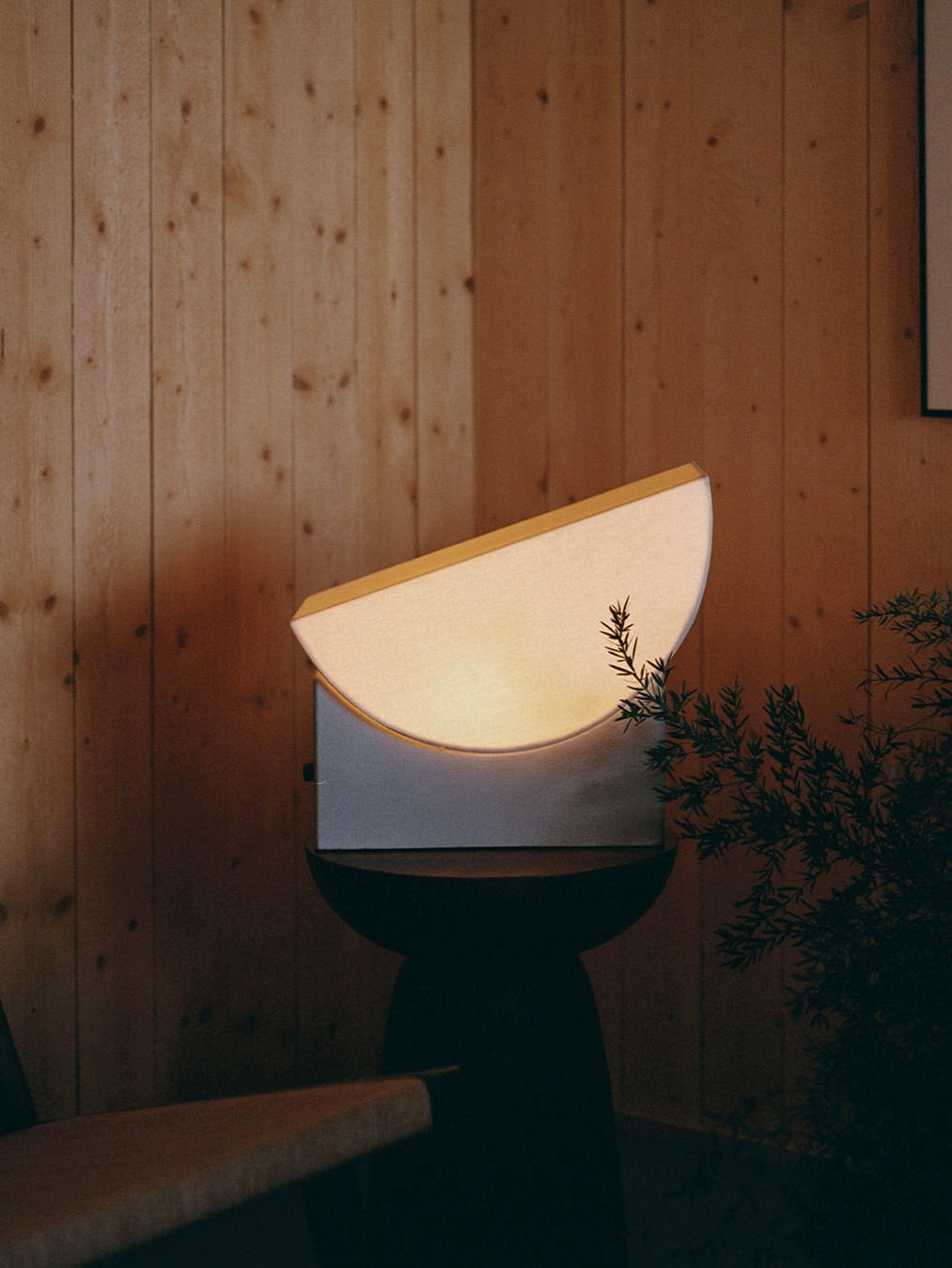 The "Reminder" Table Lamp by Enkei Lights AB, featuring a modern semicircular design, emits a warm glow in a room with wooden walls. Positioned on a small round table crafted from waste ceramics and accompanied by lush greenery, it fosters a cozy atmosphere.