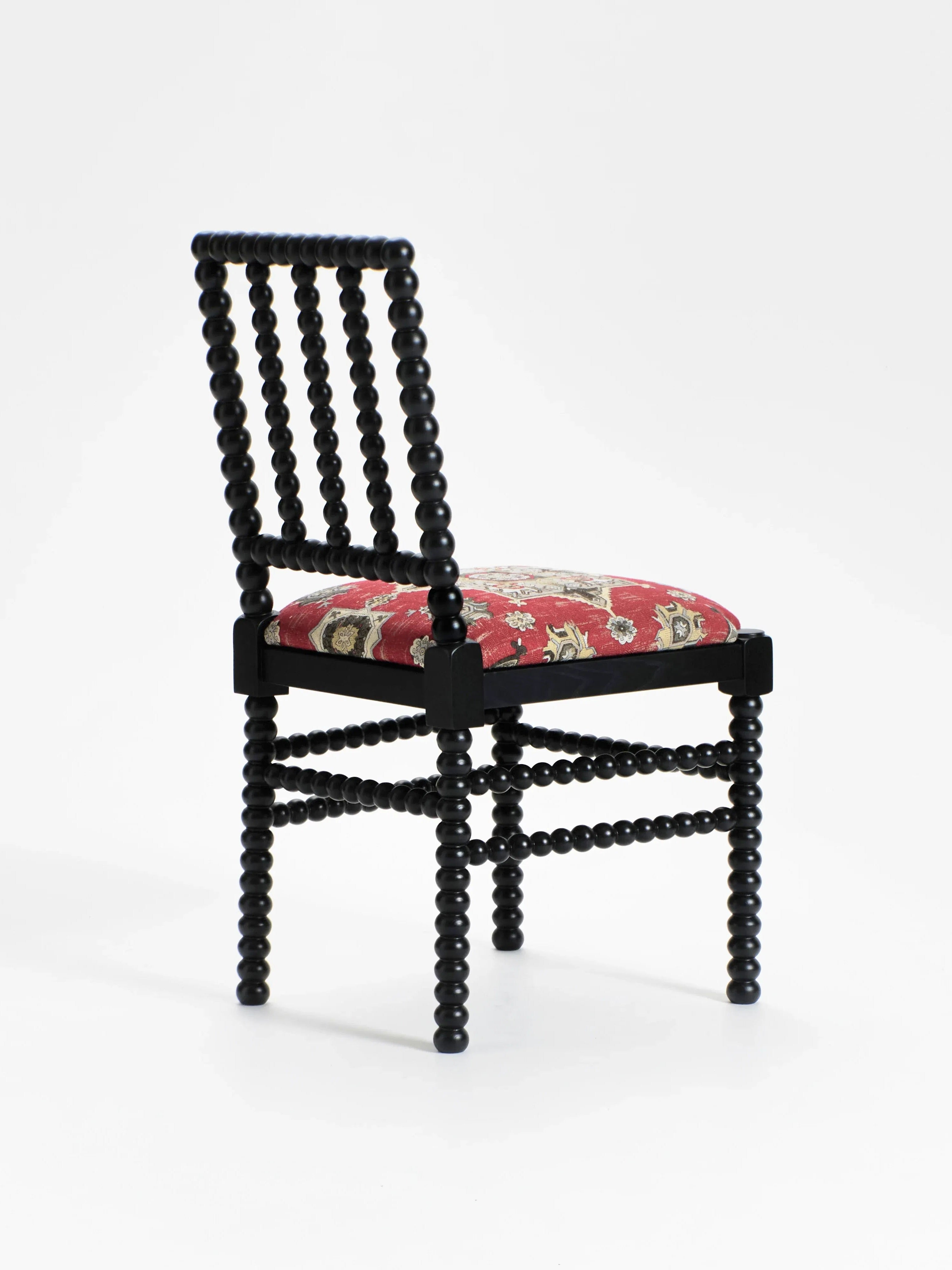 The "Bolha" Chair by Project 213A is a black wooden piece featuring ornate spindle-style legs and elegant wood-turned detailing, with a red upholstered seat showcasing an intricate floral pattern on a pristine white background.