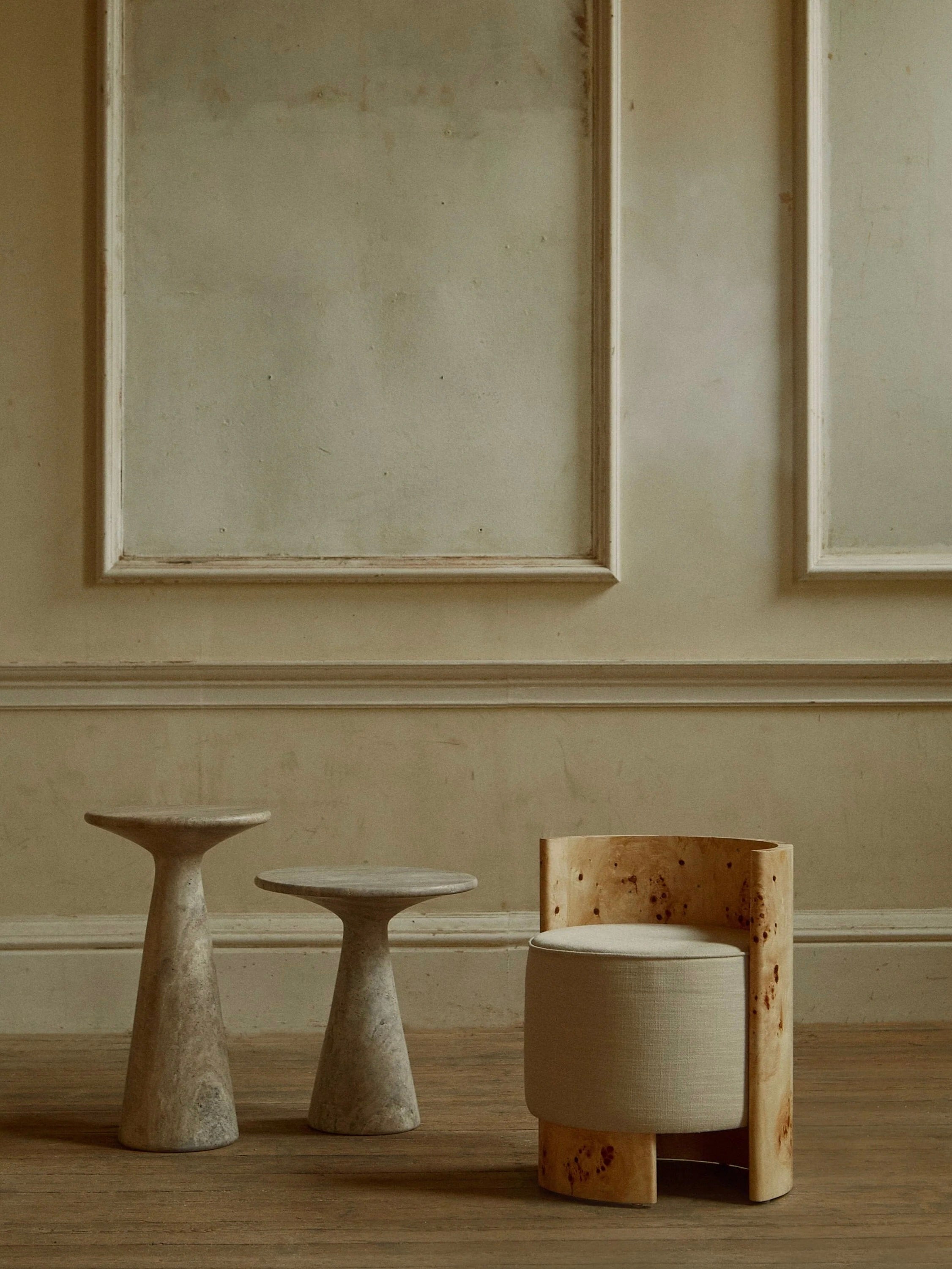 A minimalist interior showcases two sculptural stools complemented by the "Livon" Burl Armchair from Ransom & Dunn, featuring an ash burl wood frame and a beige cushion. The earthy tones and textured walls contribute to a serene atmosphere.