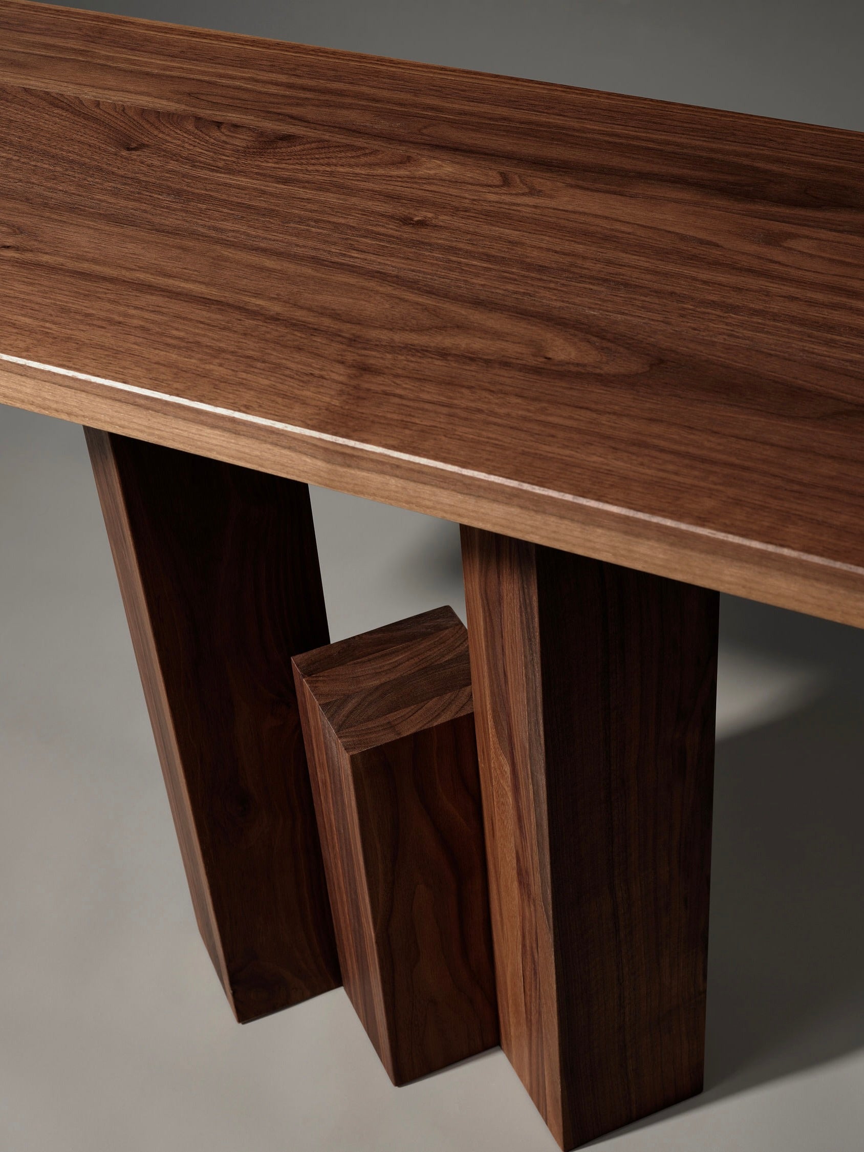 The Mokko Fenestra Console boasts a customizable design with a rectangular, polished wooden top and three varying-height block legs. Made from rich, dark wood showcasing visible grain patterns, it offers understated elegance against a plain background.