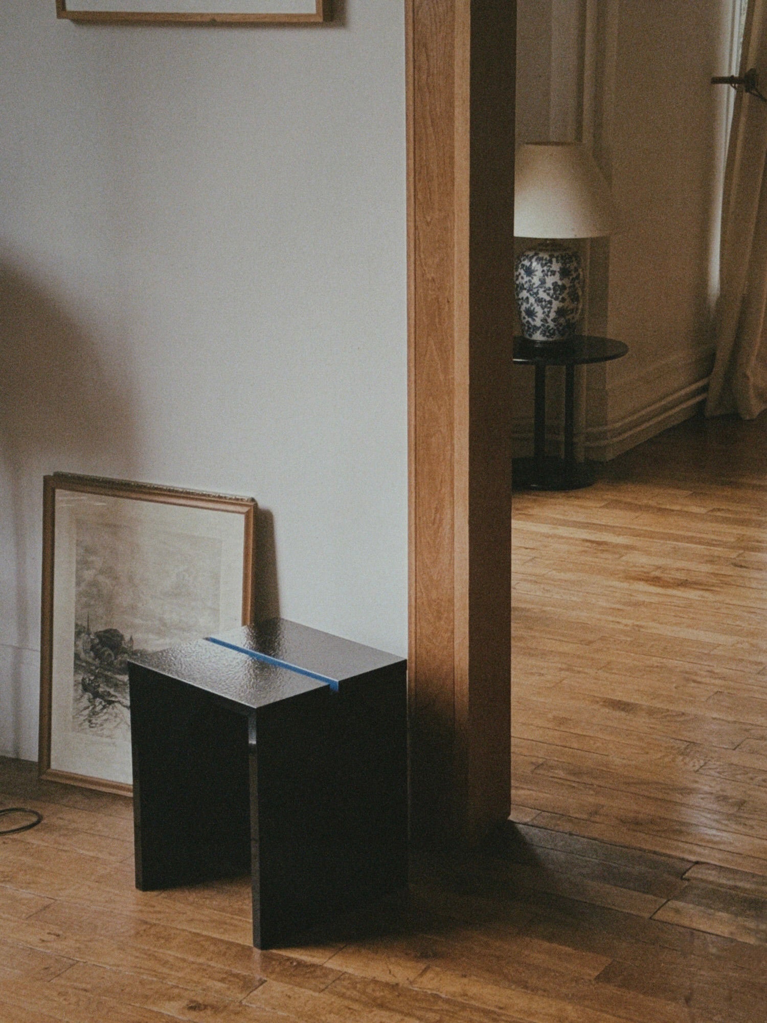 A dimly lit room with a wooden floor features a TEGET Mara Gap Stool in wet black. A framed picture leans against the white wall. In the background, a lamp with a floral patterned shade rests on laminated furniture near a window with curtains.