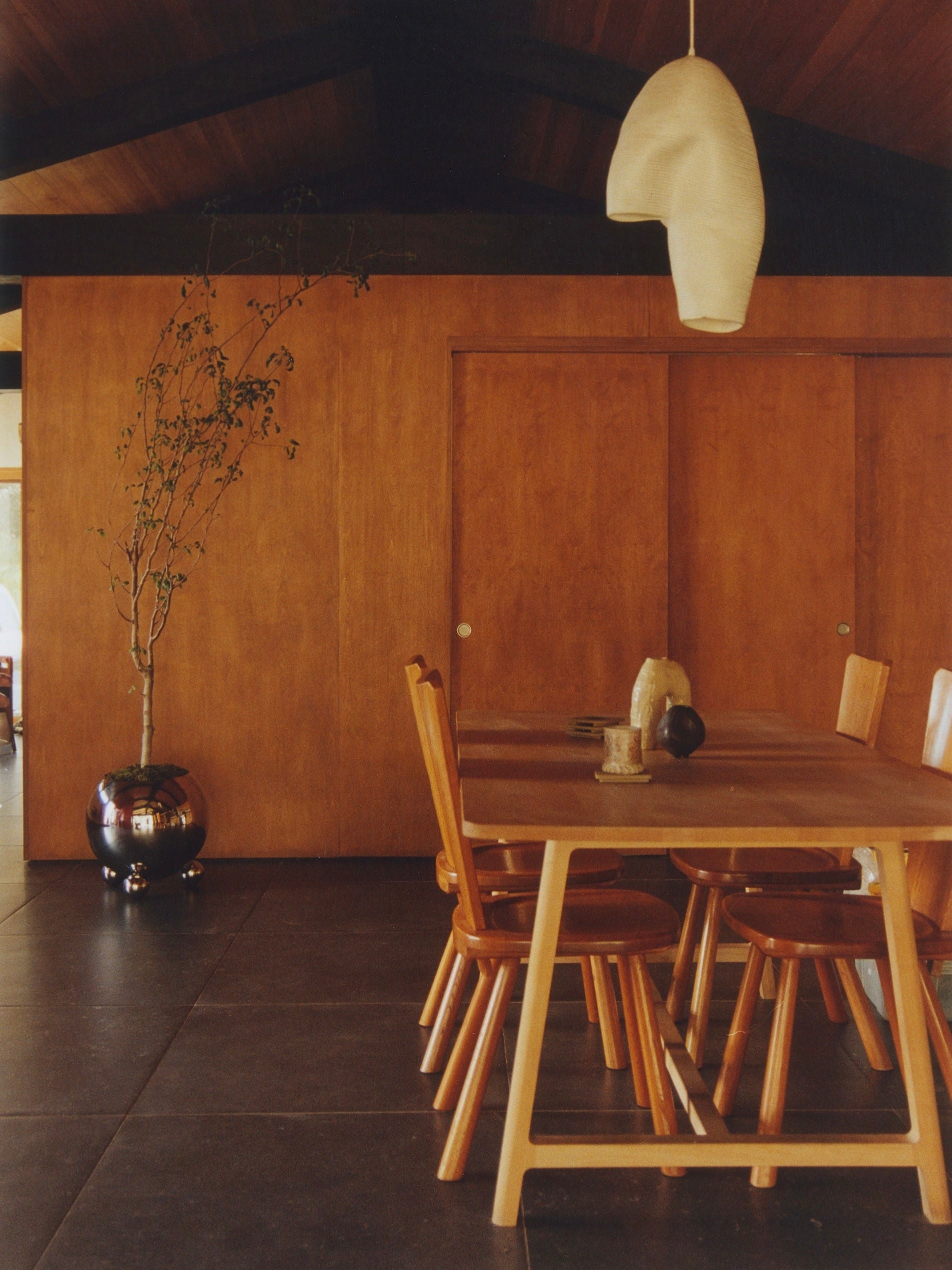 A modern dining room with wooden walls and ceiling features a long wooden table surrounded by matching wooden chairs. A uniquely shaped white pendant light hangs overhead. A potted plant stands in a large Selene Planter by MUHLY beside the table, adding a touch of greenery.