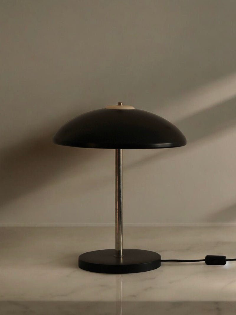 The Mushroom Table Lamp from Boga Avante Shop features a minimalist design with a sleek black dome-shaped shade and a polished chrome stem, all anchored by a flat black circular base. Perfect for mid-century modern interiors, this stylish lamp is showcased on a marble surface against a light-colored backdrop, complete with its attached black power cord.