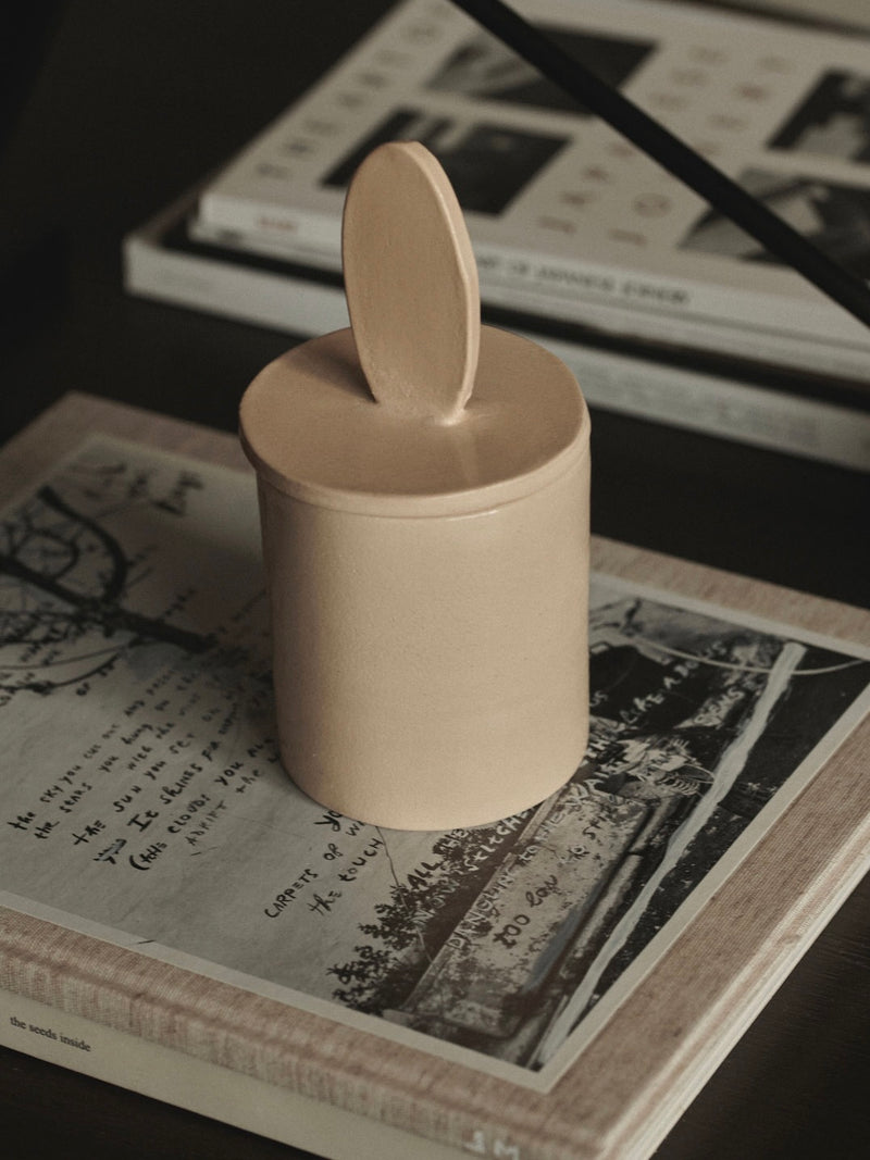 A tan, lidded ceramic jar with an oval handle sits atop an open coffee table book featuring black and white imagery and handwritten text. This limited edition piece exudes elegance—a nod to the artisanal 2222Studio Sculptural Scented Candle glimpsed in the background.