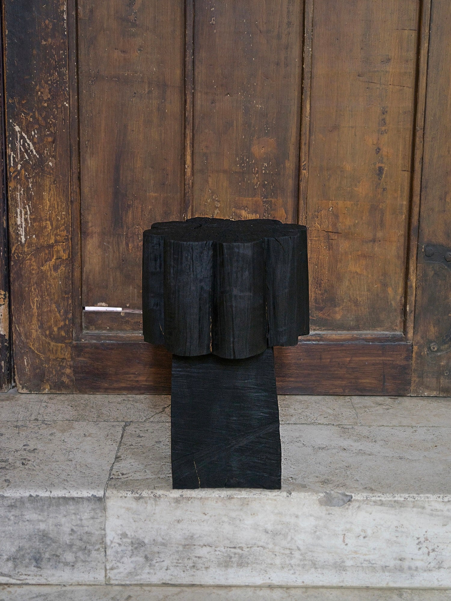 A Burnt Black Coffee Table/Stool by Touch With Eyes, featuring a scalloped edge and textured finish, stands on a stone surface in front of a weathered wooden door.