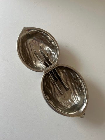 The septembre studios Nutcracker—small, metal, and clam-shaped—opened to reveal several screws inside, evoking a practical nutcracker design and resting on a white surface.